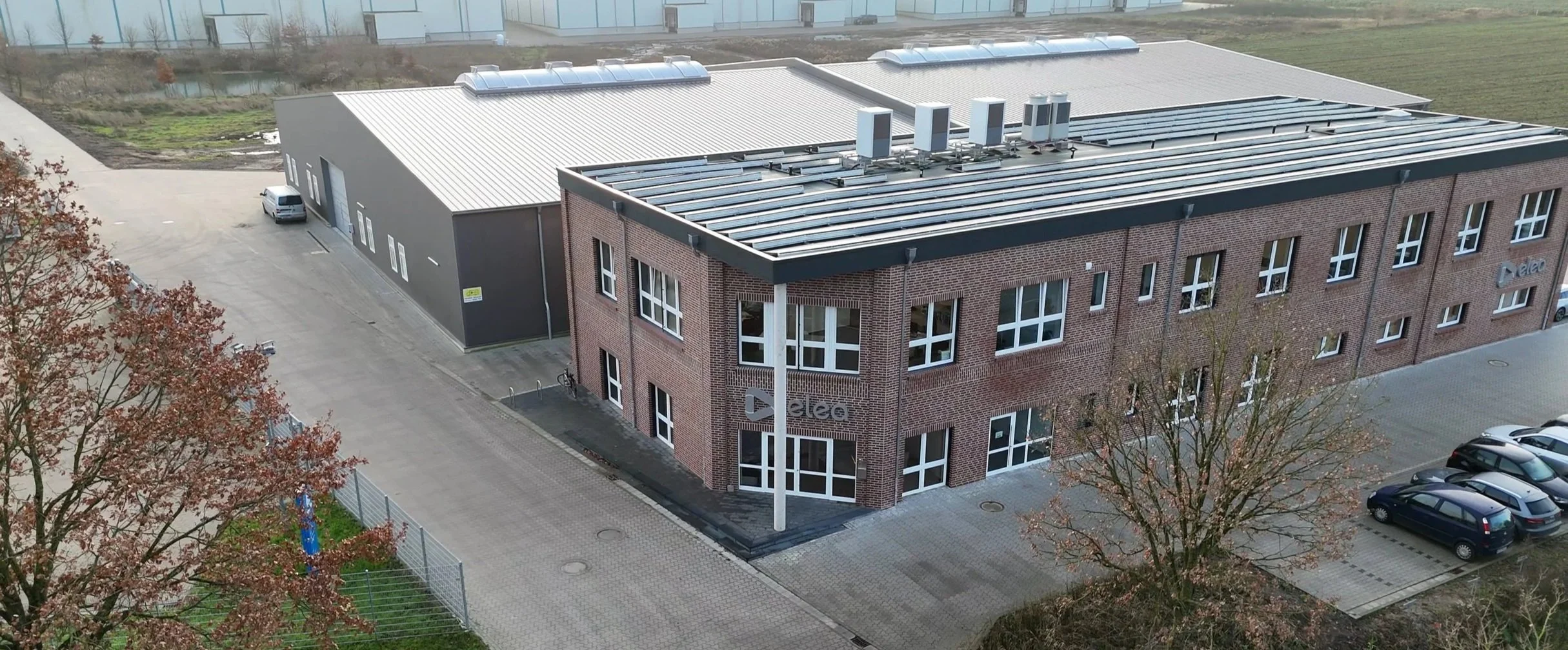 An aerial view of a modern office building with brick exterior, large windows, and solar panels on the roof. There is a parking lot with several cars, trees in the foreground, and a neighboring industrial building.