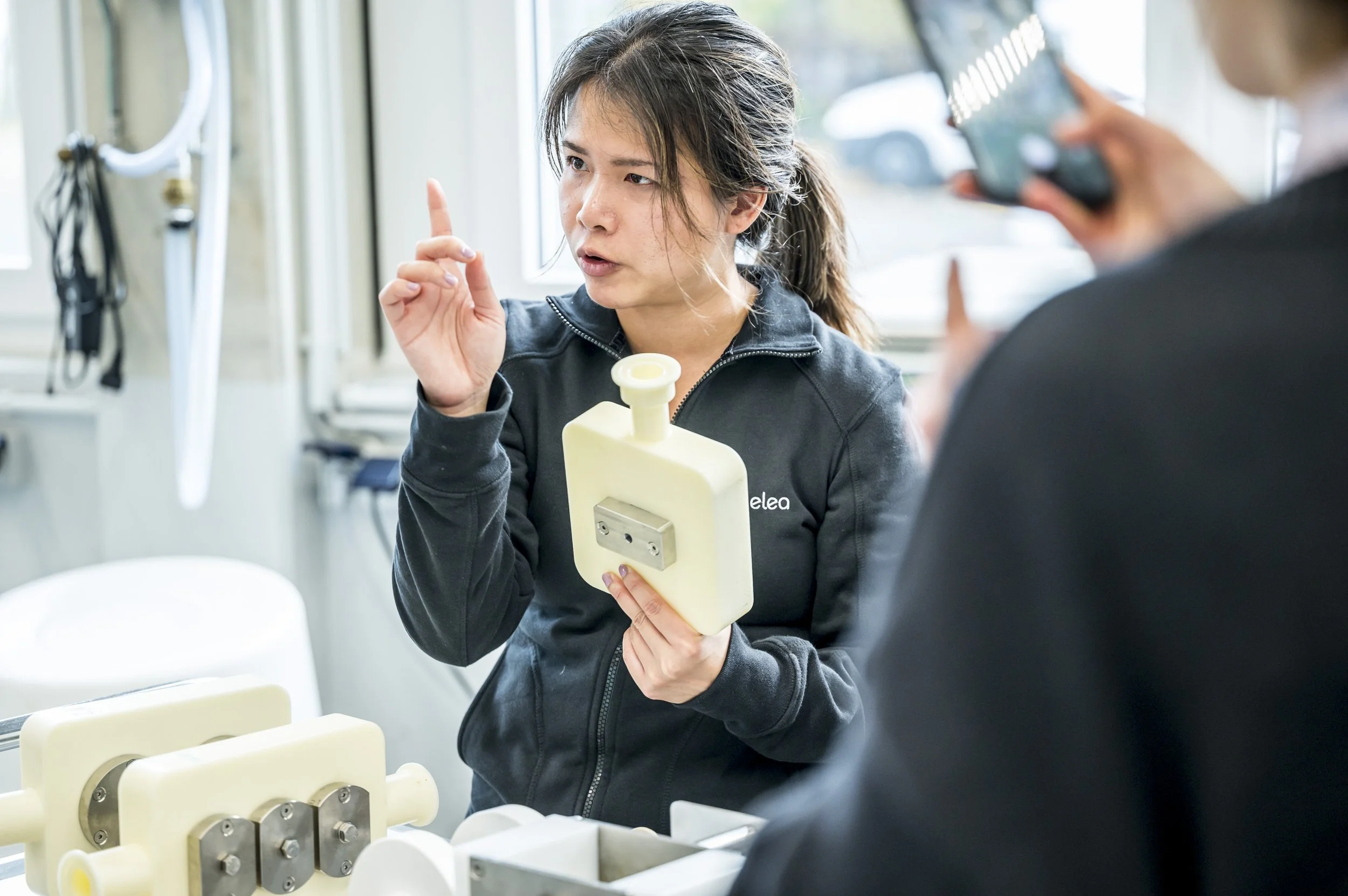 Woman in black jacket demonstrating a medical device to another person in a clinical setting.