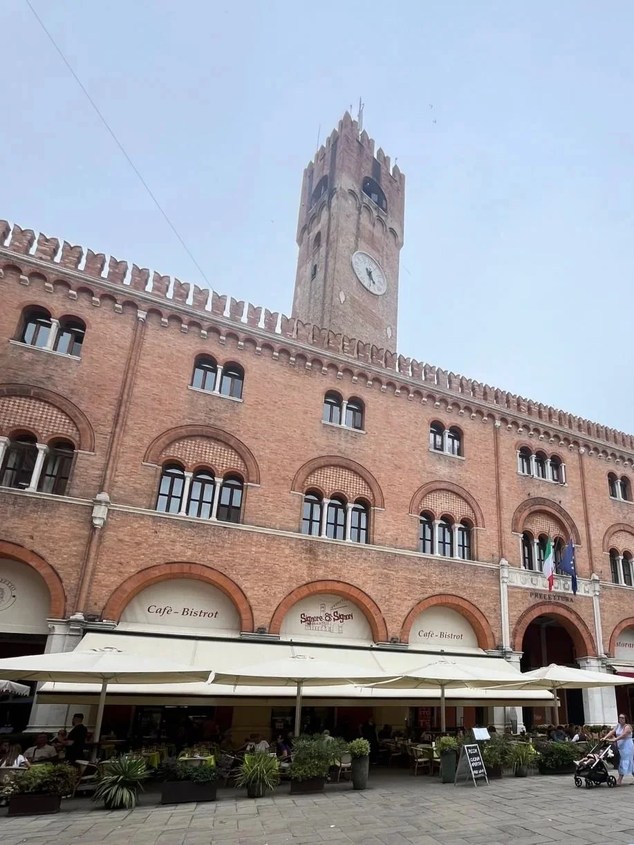 Historic brick building with a clock tower, outdoor cafe seating, shops, and flags in front, in a city square.