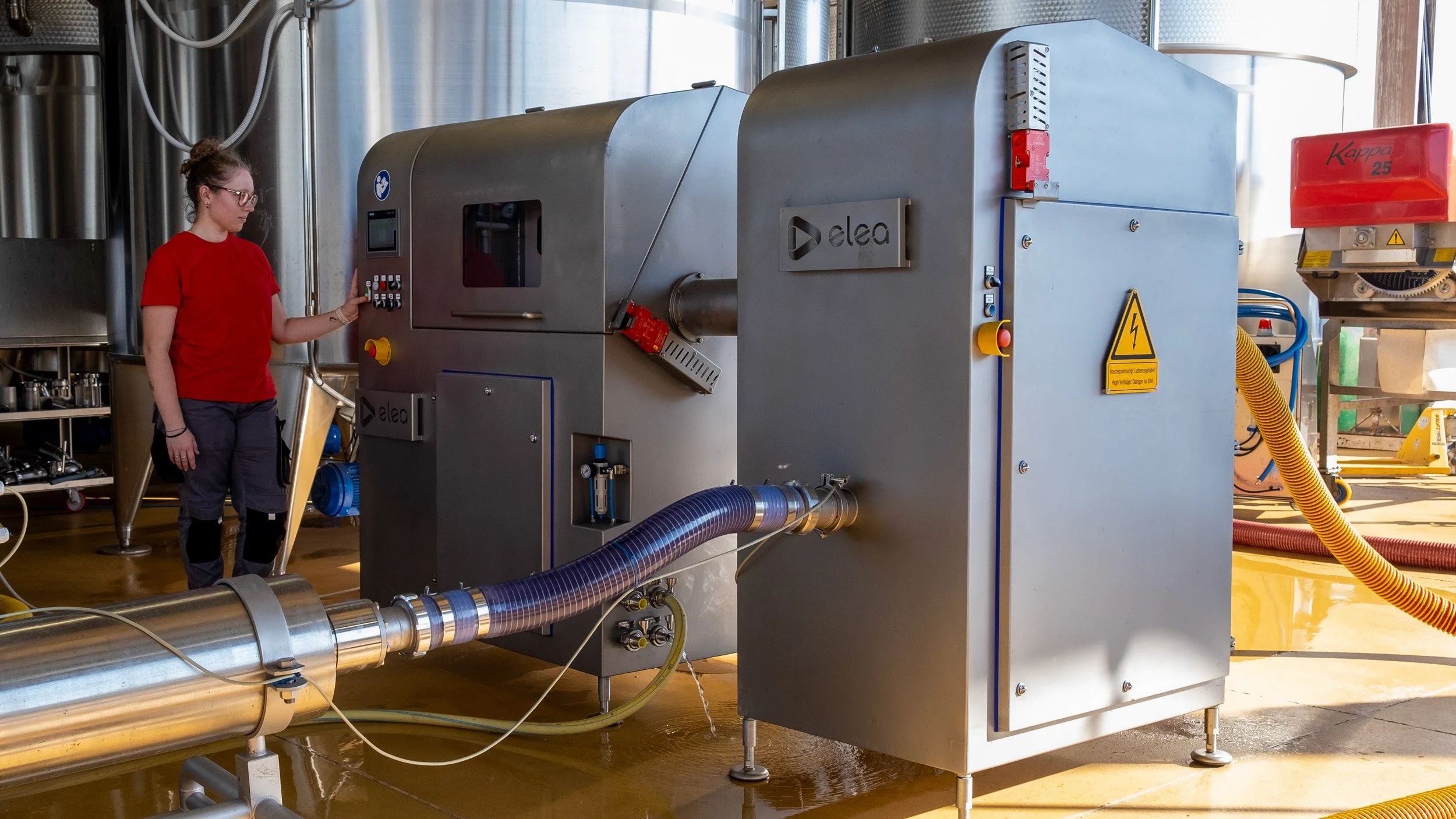 A woman in a red shirt operating industrial equipment with controls in a factory or plant setting, with stainless steel machinery and yellow hoses around.