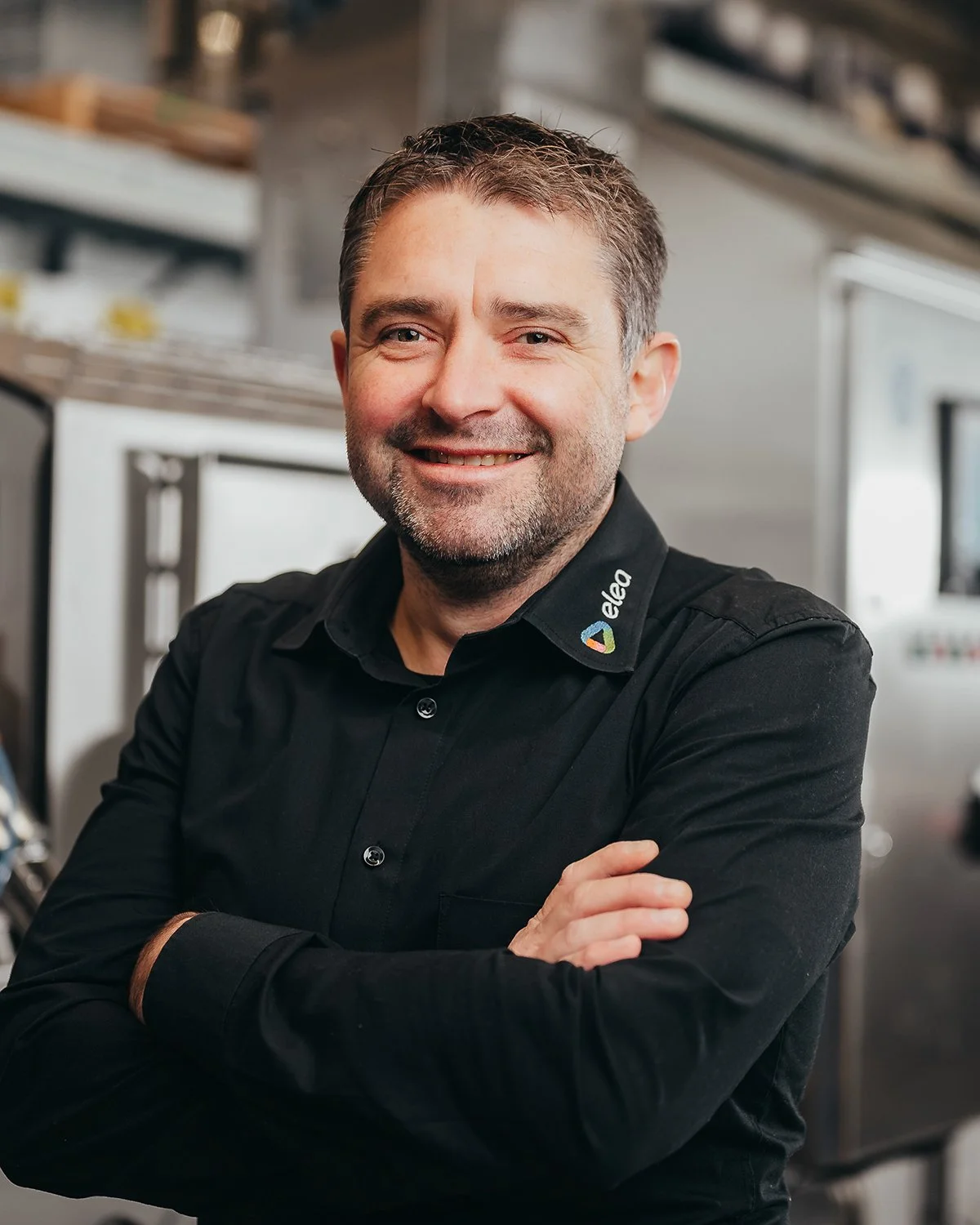 Smiling man in black shirt with "elea" logo, standing indoors with arms crossed.