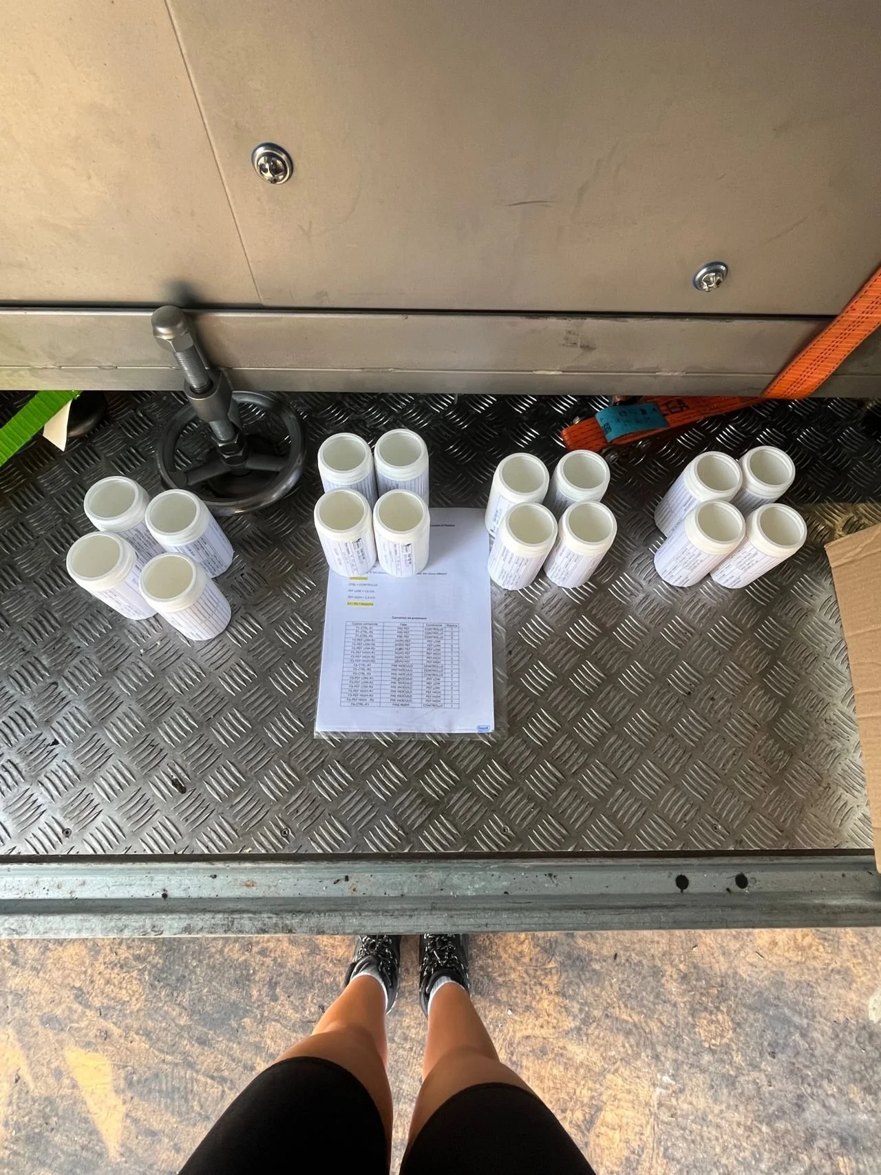 A person standing in front of a metal counter with several white cylindrical containers and a clipboard with a chart on it, on a textured metal floor.
