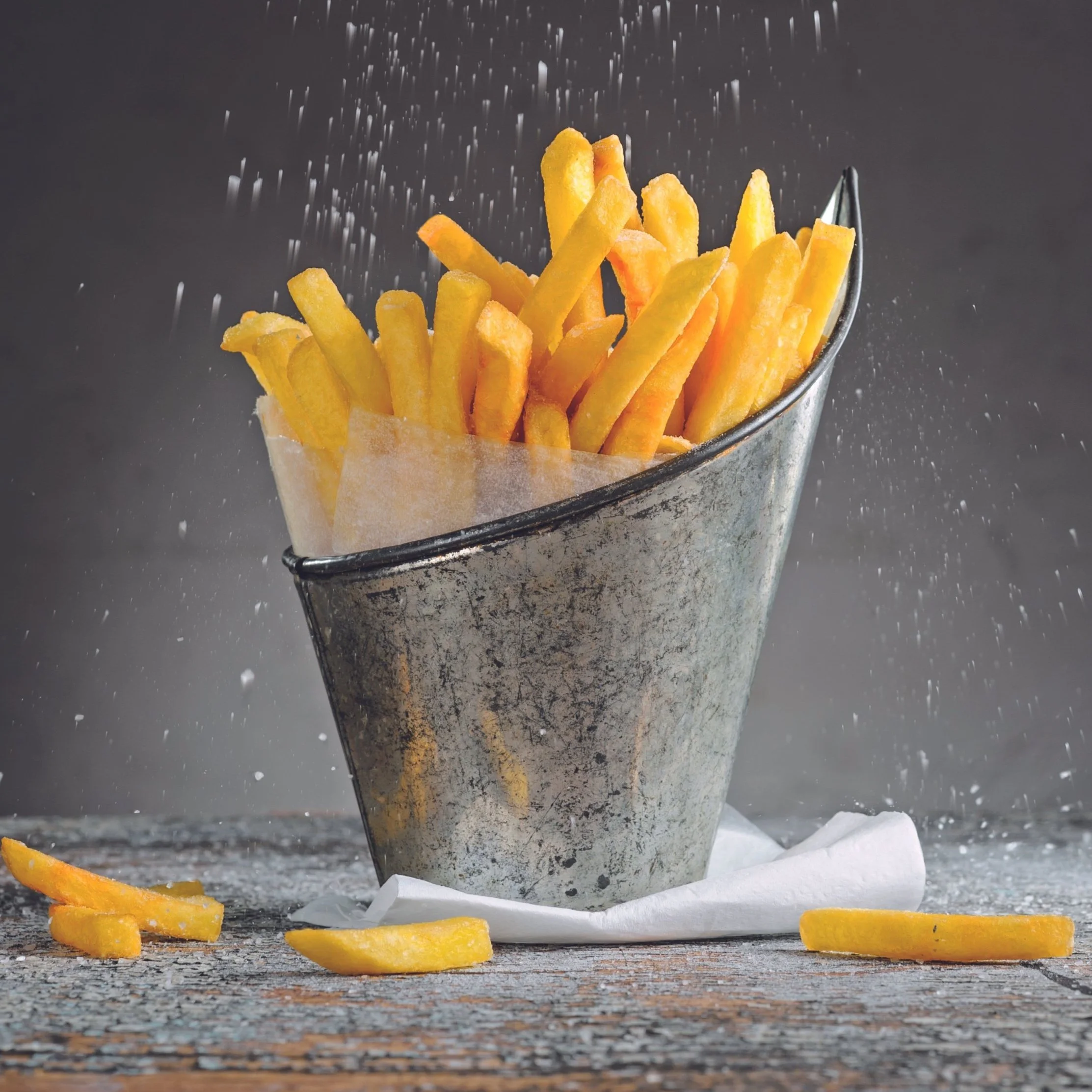 French fries in a metal bucket with salt being sprinkled.