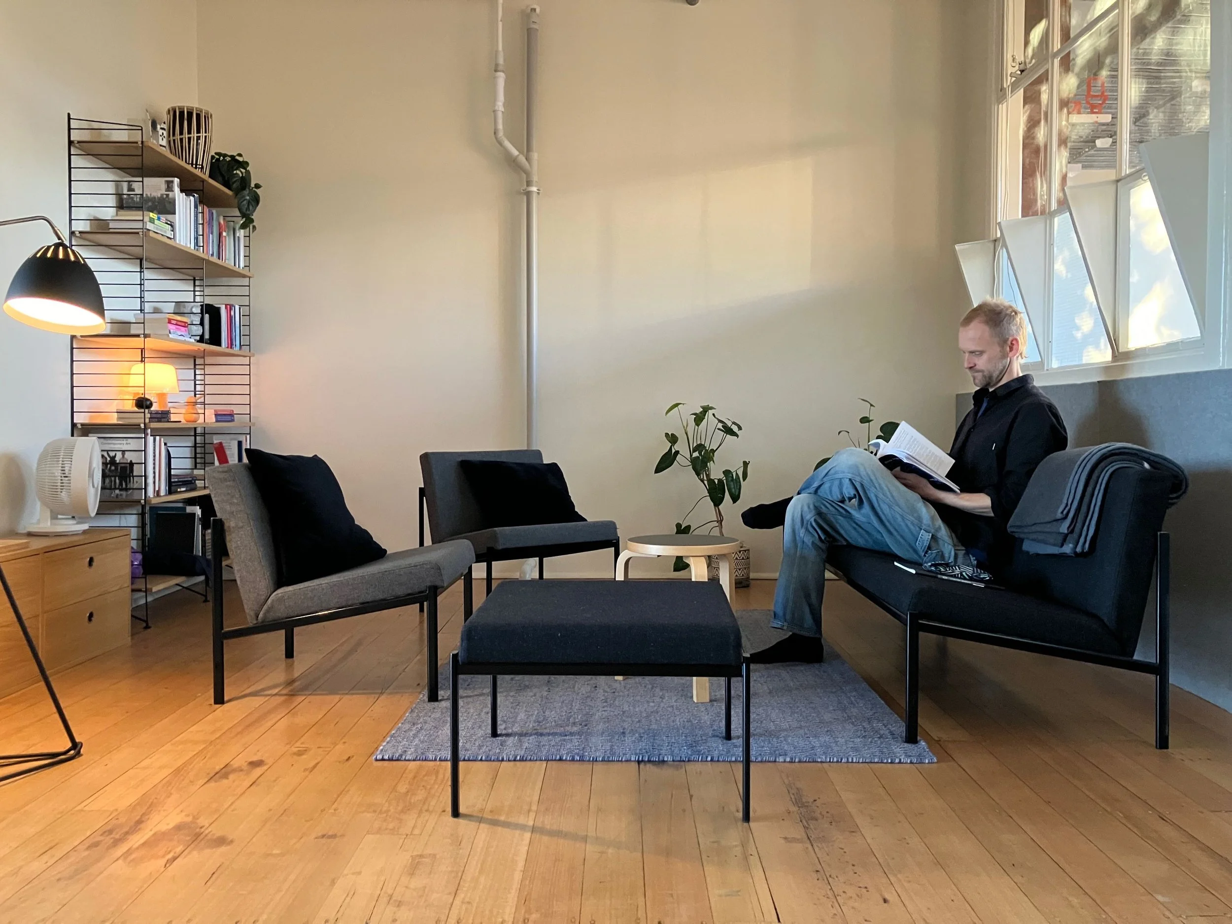 Daniel Costin reading in his studio at Collingwood Yards — seated by a window in a warm, naturally lit space with dark armchairs, a bookshelf, and a small plant.