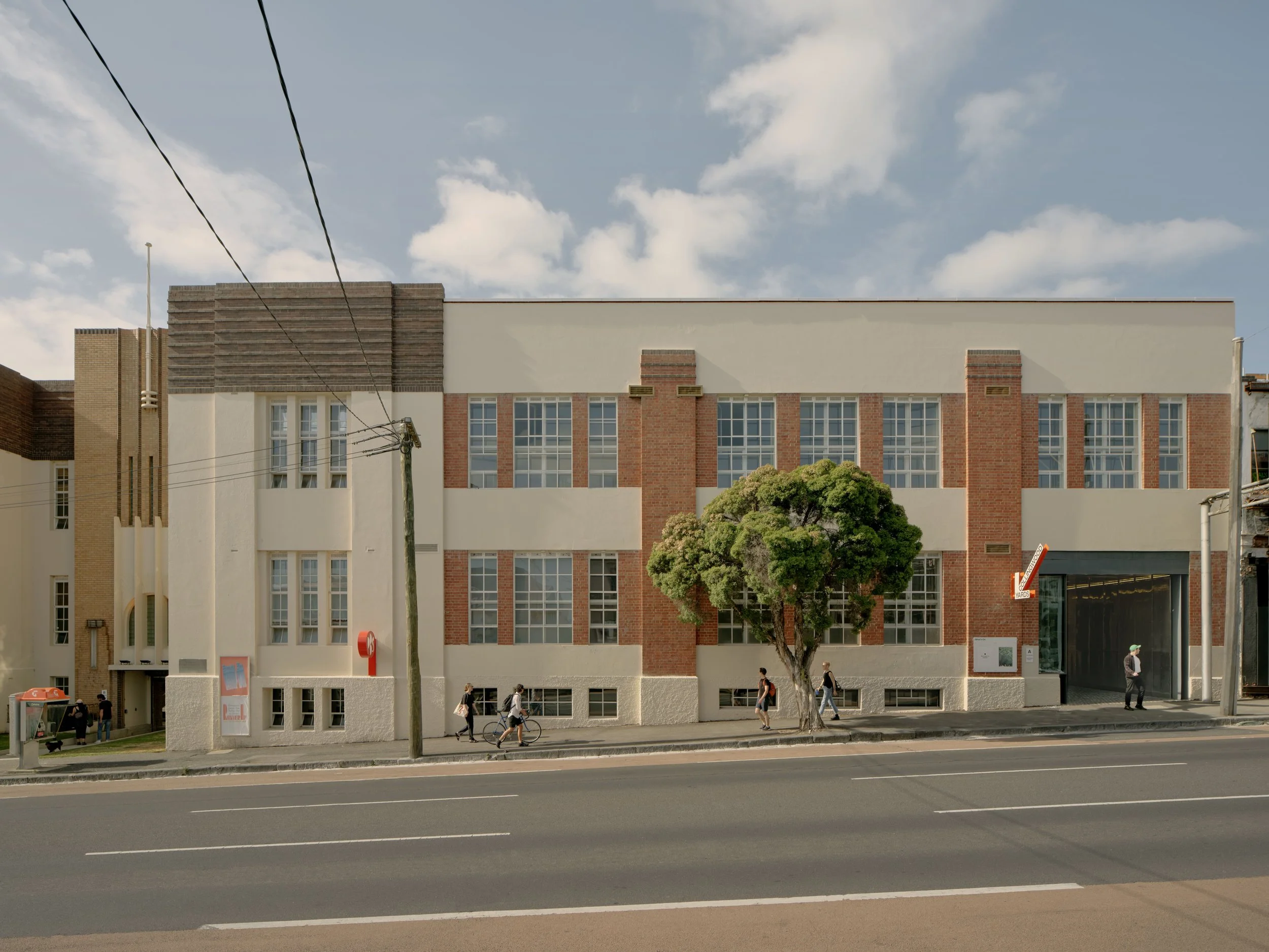 The street frontage of the Johnston Street Building at Collingwood Yards — a cream heritage façade with red-brick detailing and tall windows, a leafy tree and people passing on the footpath below.