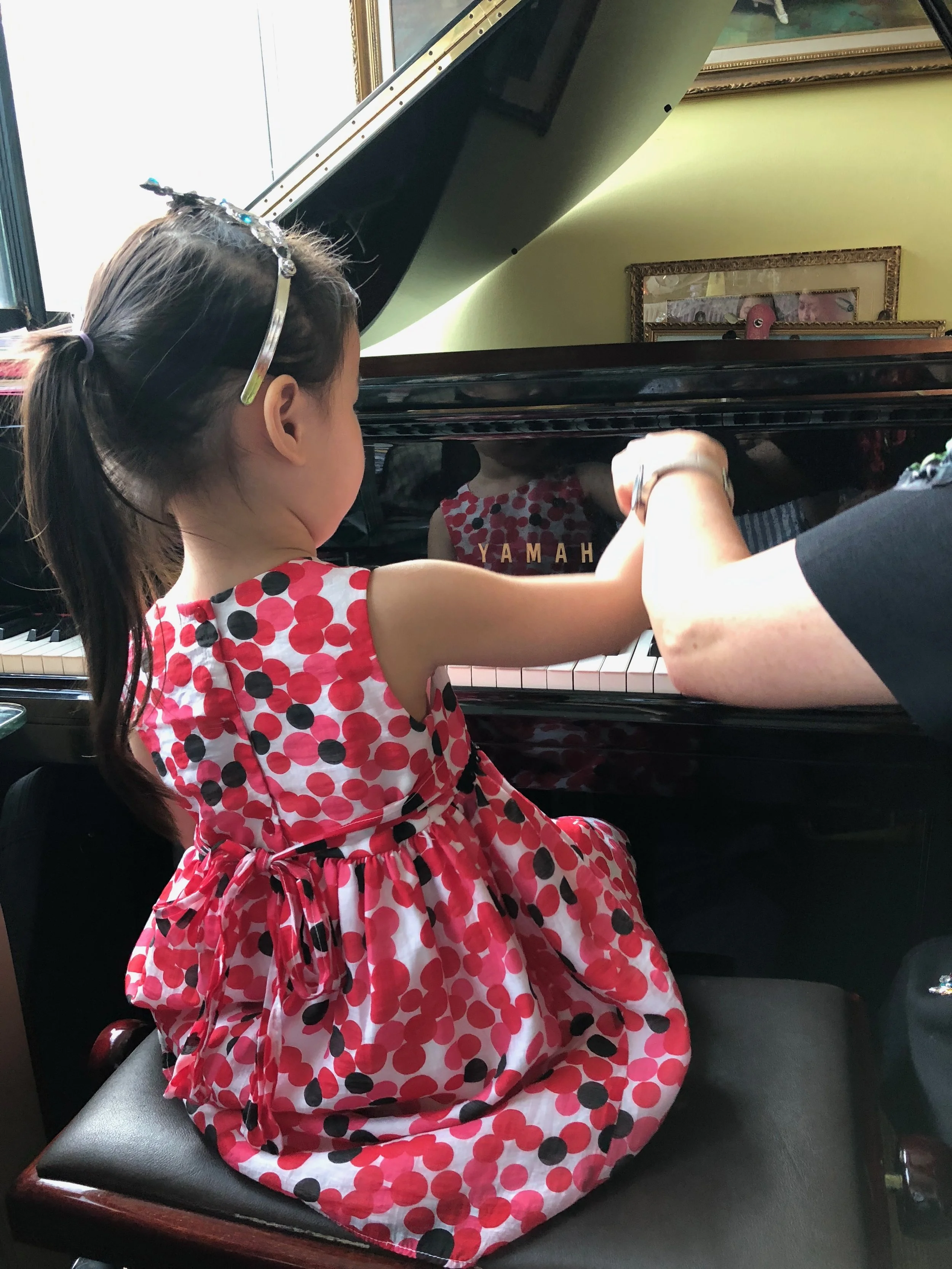 A young girl with a ponytail, wearing a red and white polka dot dress, is sitting at a piano, reaching with her left hand to press the keys.