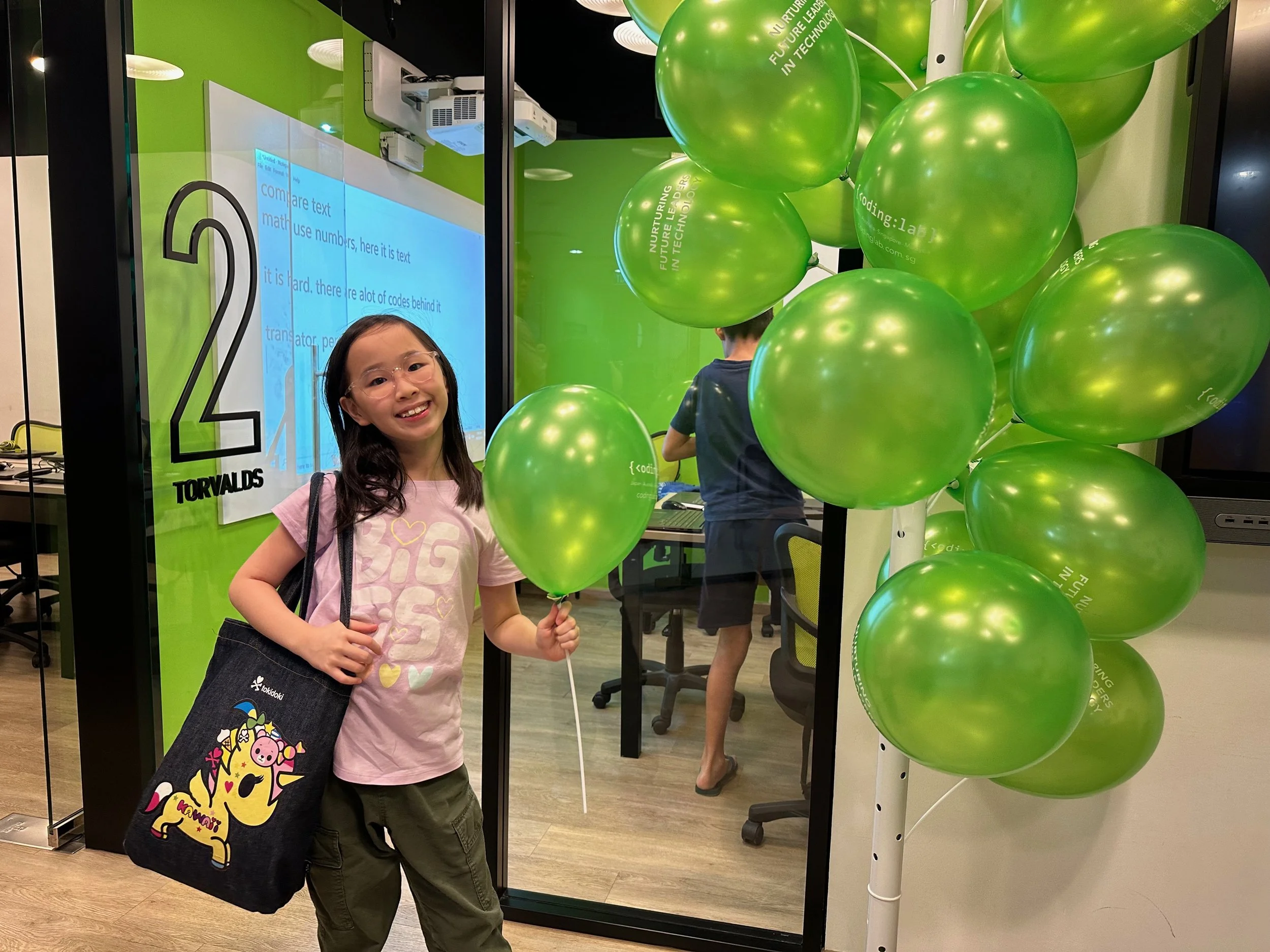A smiling young girl wearing glasses holding a black tote bag with a colorful unicorn illustration, standing in front of green balloons at an indoor tech event.