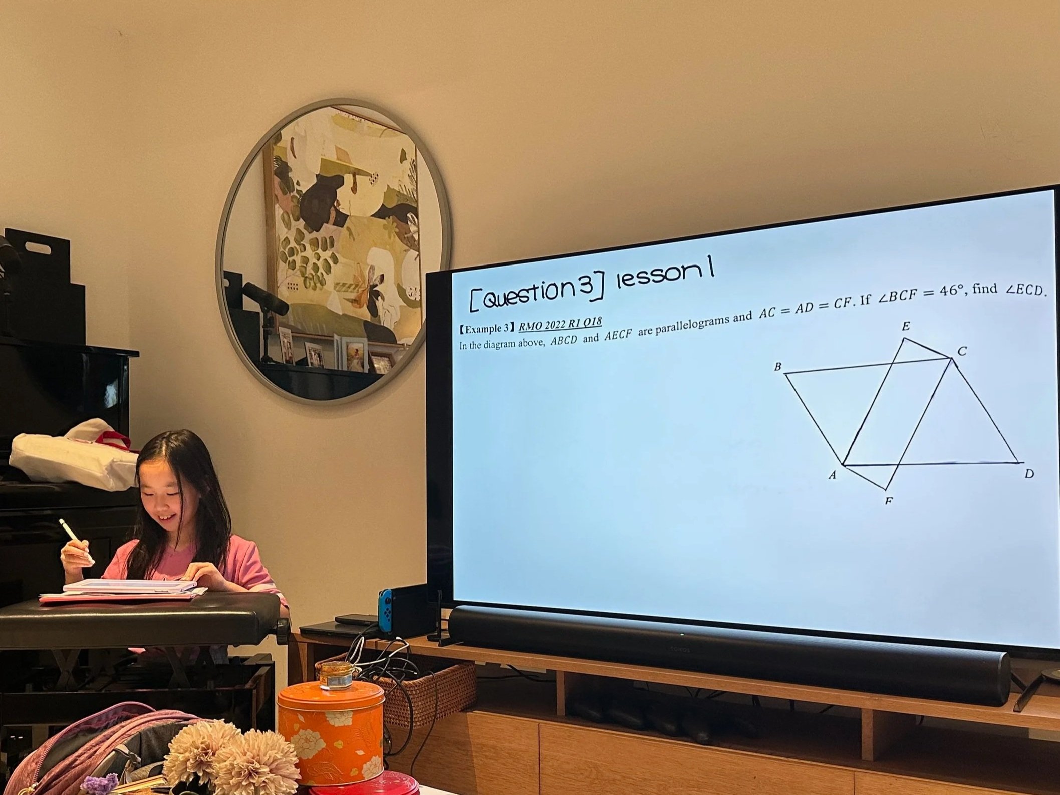 A classroom or study area with a large digital screen displaying a geometry problem about parallelograms and a young girl sitting at a keyboard, smiling while holding writing instruments and a notebook.