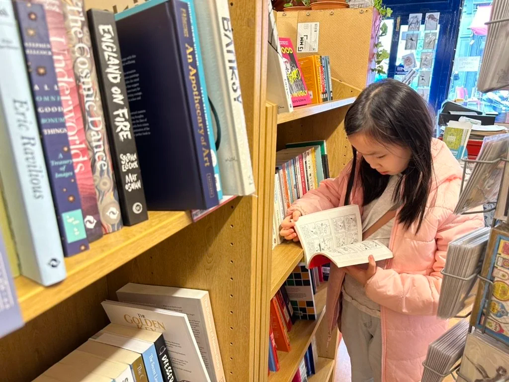 A young girl in a pink jacket and gray pants browsing and reading a comic book in a bookstore aisle.