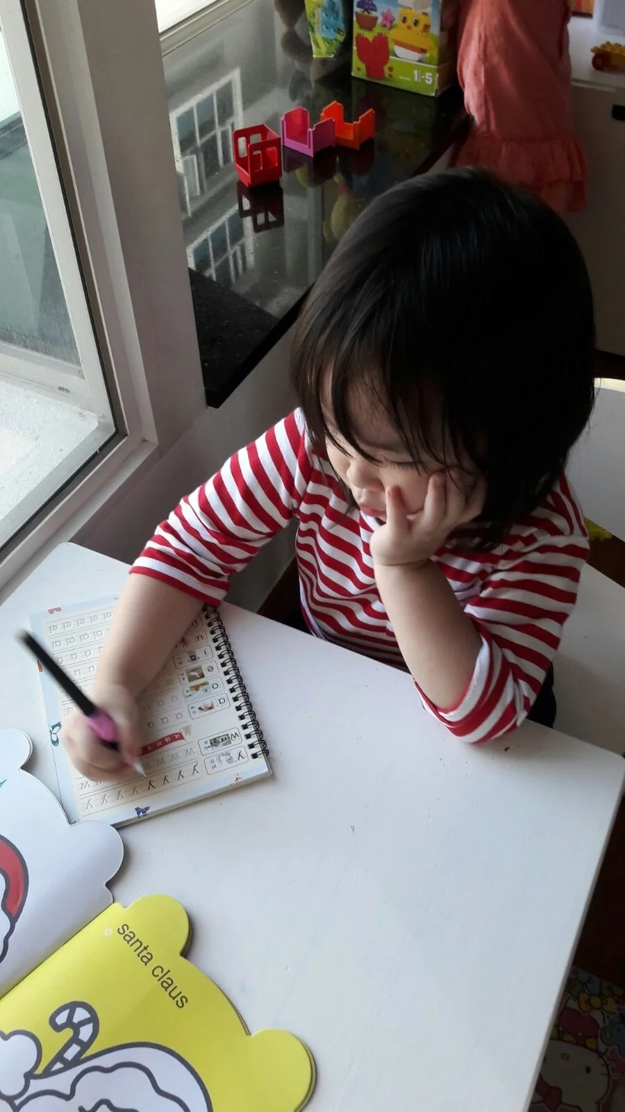 A young child with dark hair and wearing a red and white striped shirt is sitting at a white table, doing a homework or activity in a workbook with a black marker. The child has one hand on their face, appearing focused or thoughtful. There are holiday-themed cutouts on the table, including one yellow with a candy cane and the label 'santa claus.' In the background, a window shows an outdoor view, and a countertop holds colorful toys and a box with animal illustrations.