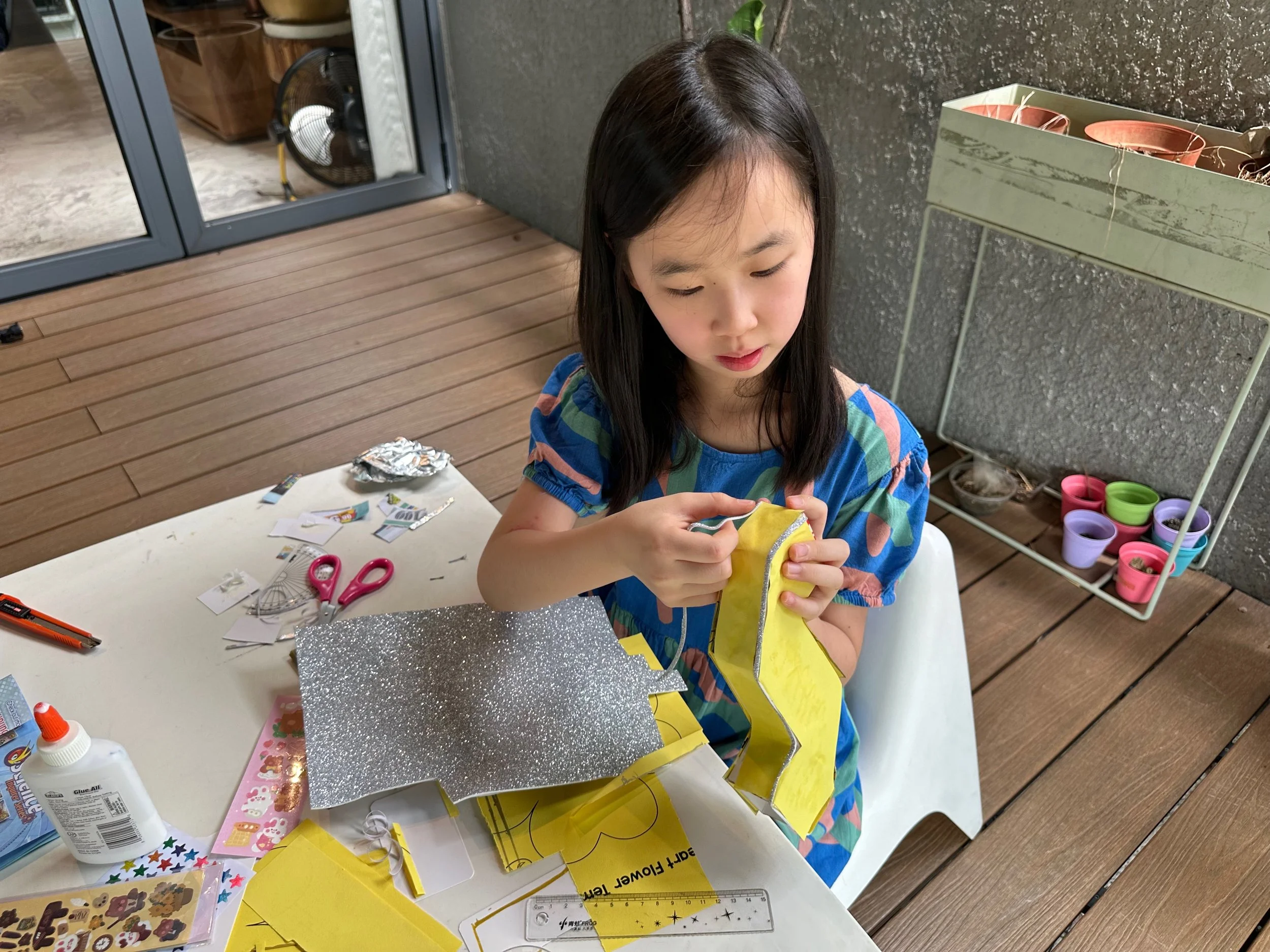 A young girl with long dark hair wearing a blue, green, pink, and purple striped shirt sits at a white table, holding yellow paper and working on a craft project. The table has various craft supplies including scissors, glue, stickers, and decorated paper. Behind her, there is a gray textured wall, a window or glass door, and a tiered plant stand with terracotta pots.