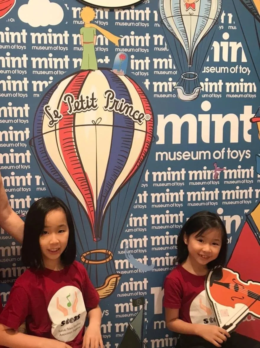 Two young girls in red shirts standing in front of a blue and white backdrop with hot air balloons and the text 'Le Petit Prince' and 'mint museum of toys' repeated repeatedly. One girl is holding a violin and smiling, while the other has her mouth slightly open.