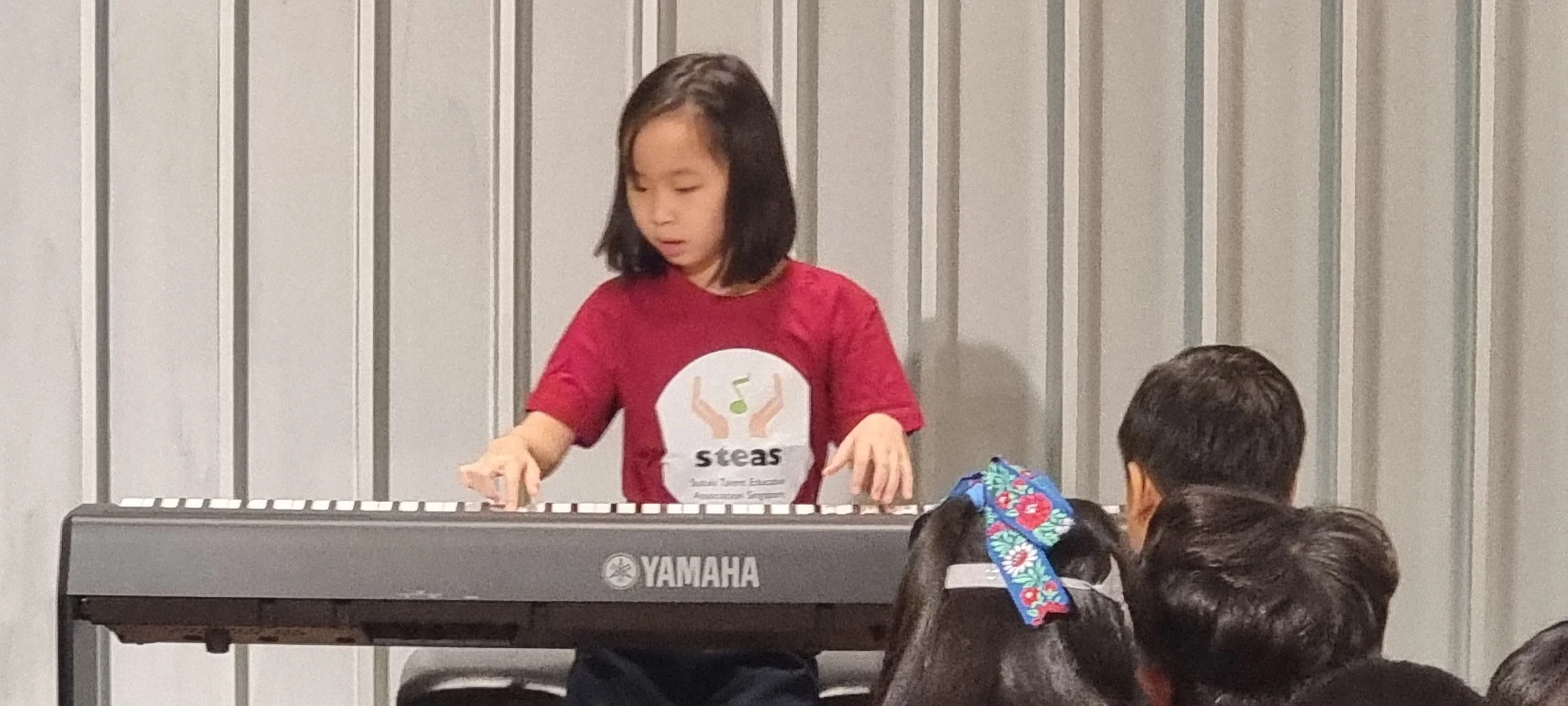 Young girl playing a Yamaha keyboard during a performance, wearing a red shirt with 'Steas' logo, in front of an audience.