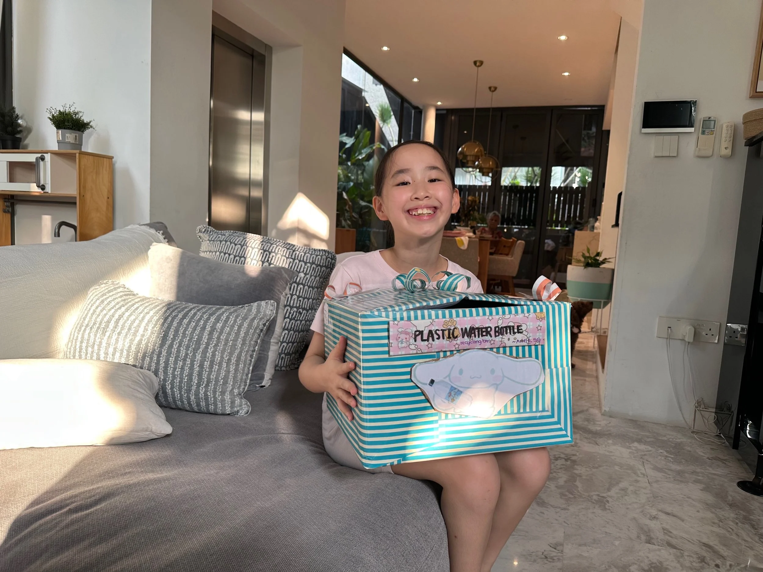 A young girl with a big smile holding a blue and white striped box labeled 'Plastic Water Bottle' while sitting on a gray couch in a modern living room.