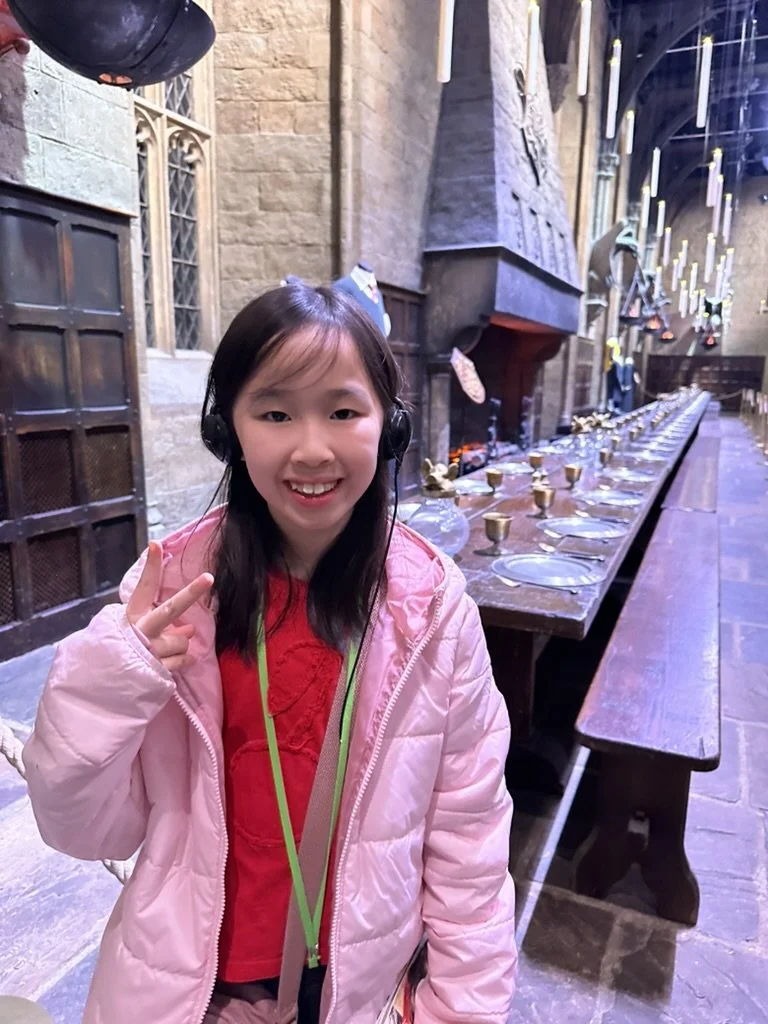Young girl smiling and making a peace sign, wearing a pink jacket and headphones, in a castle-like setting with a long dining table set with plates and goblets.