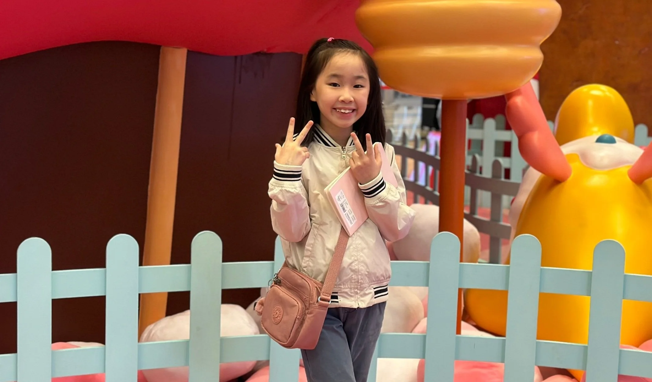 A young girl smiling and making a peace sign with both hands inside a playful, colorful playground area. She is wearing a beige jacket with black and white striped collar, carrying a pink crossbody bag with a tag, and standing in front of a light blue picket-style fence with a large yellow and pink structure in the background.