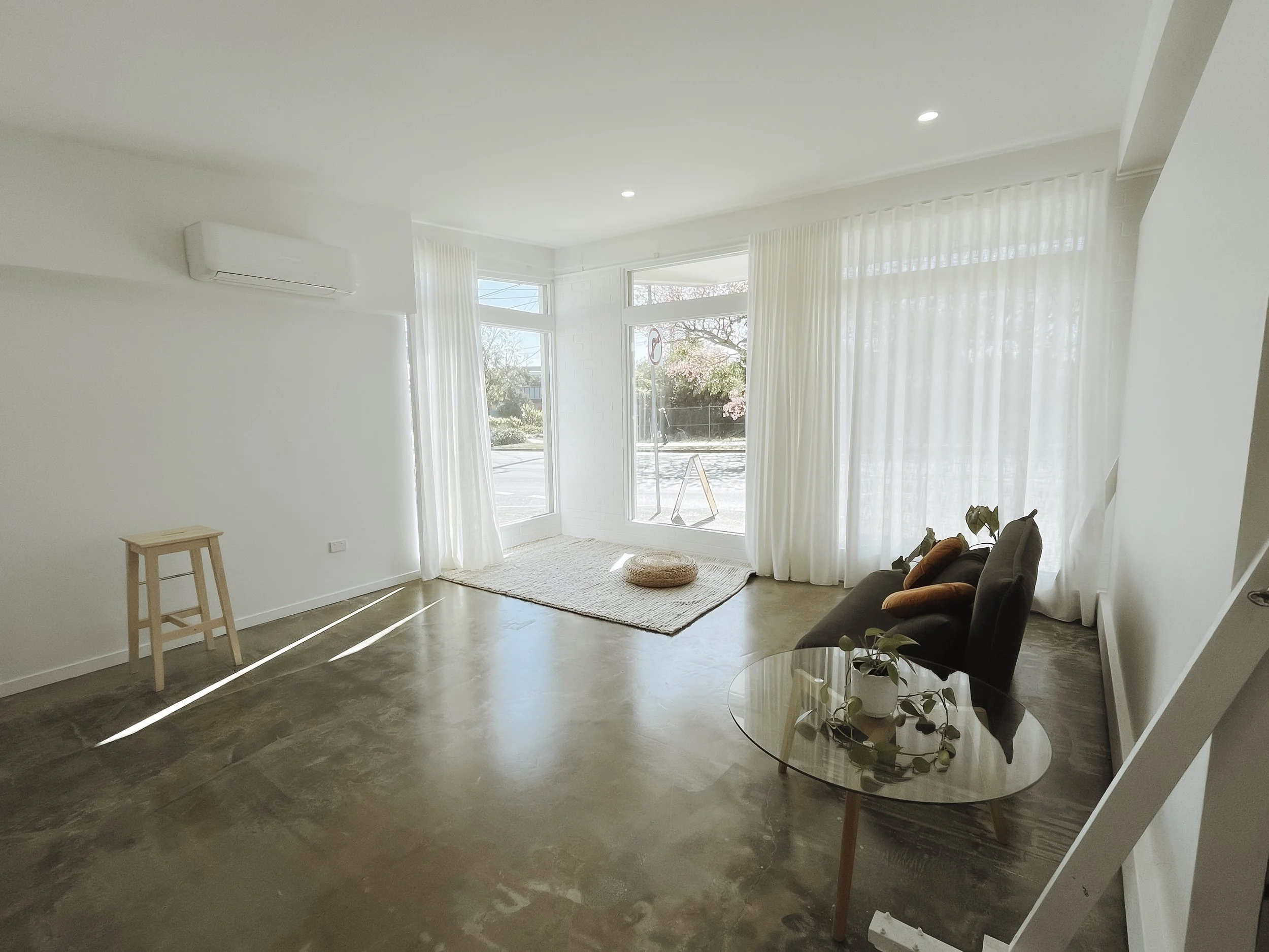 Minimalist living room with large windows, sheer white curtains, gray polished floor, black sofa, wooden stool, round glass coffee table, and potted plant.