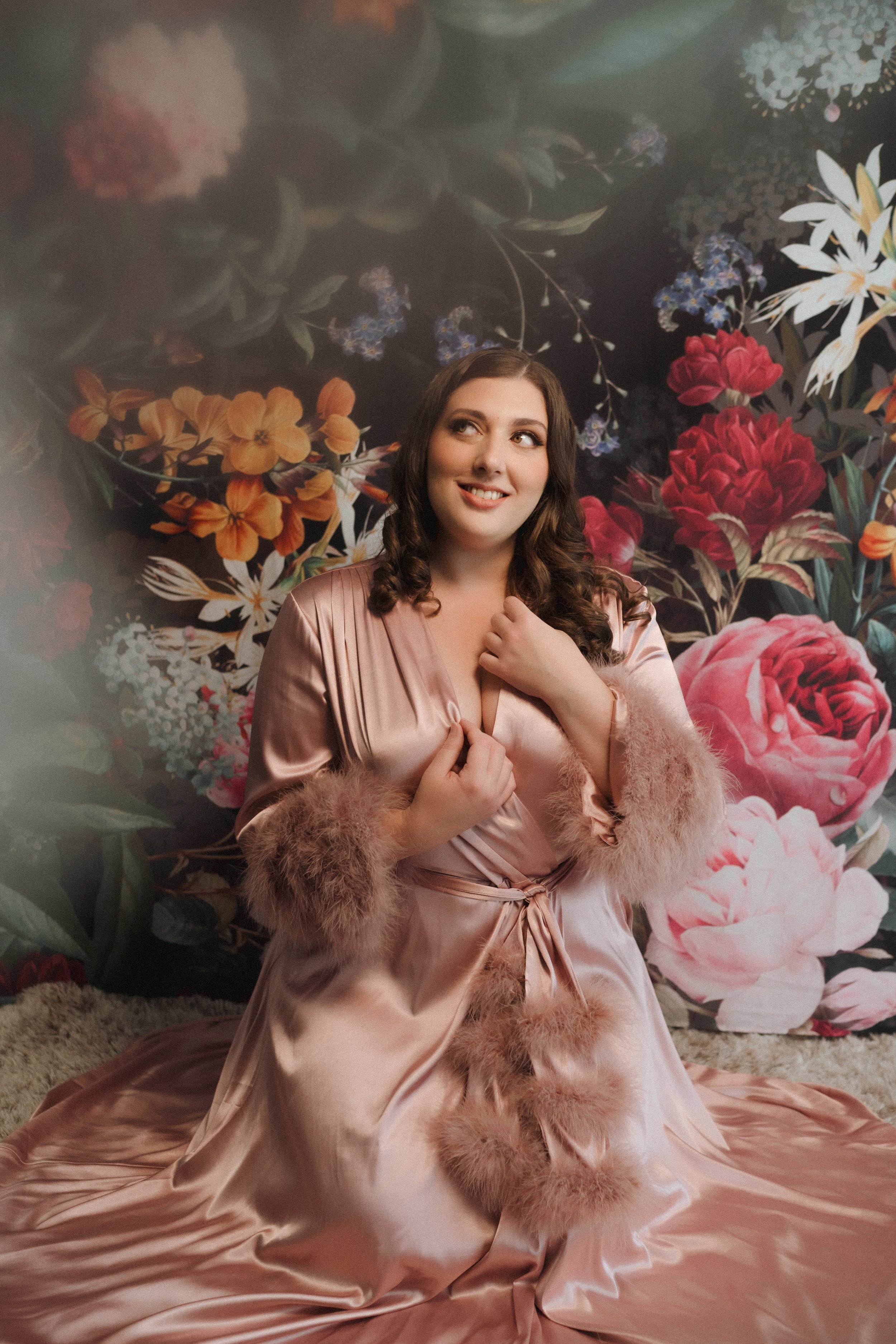 Woman in a pink satin robe with fur trim, seated against a floral backdrop, looking upwards.