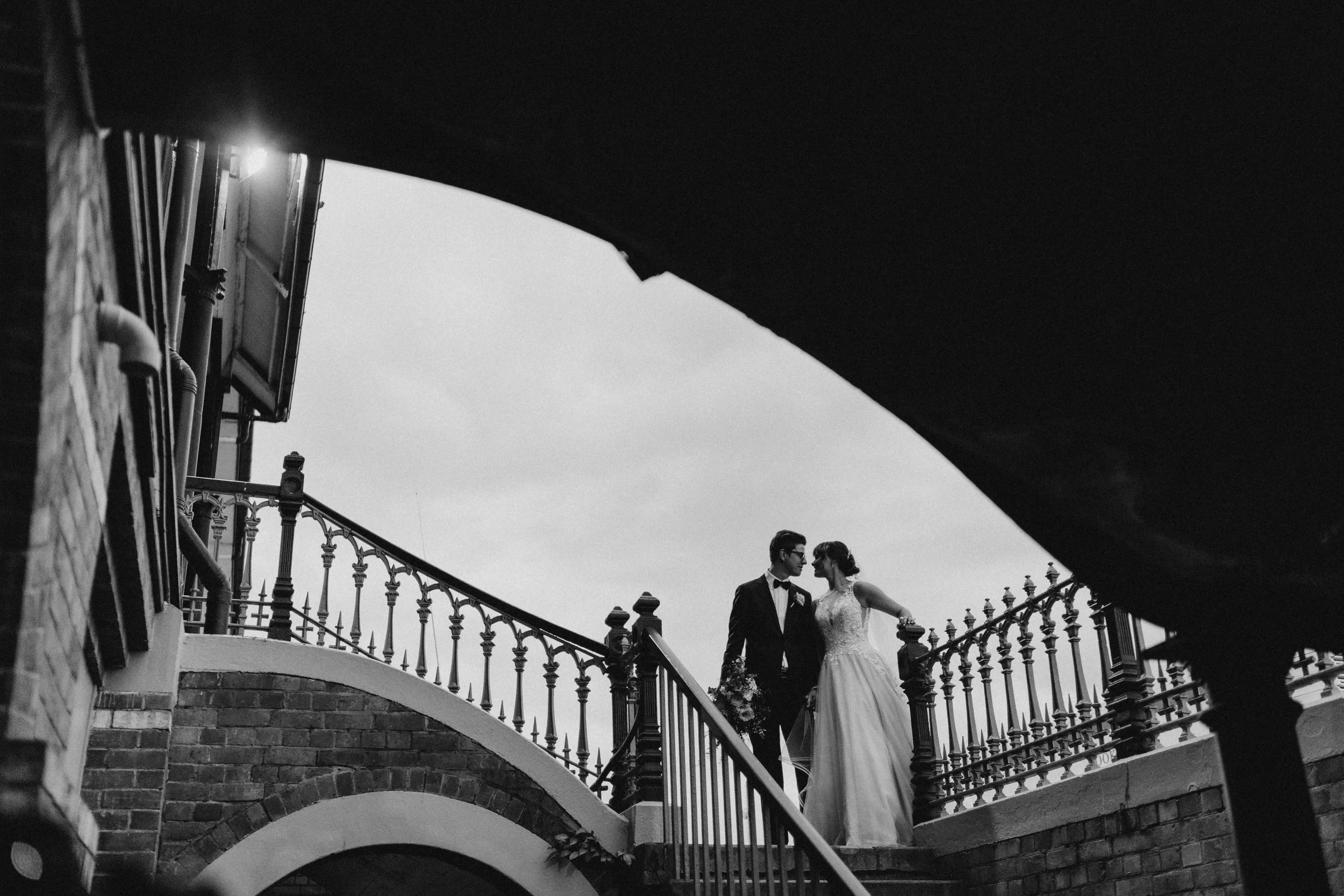 Black and white photo of a couple in wedding attire standing on a brick staircase, with ornate railing and arches.