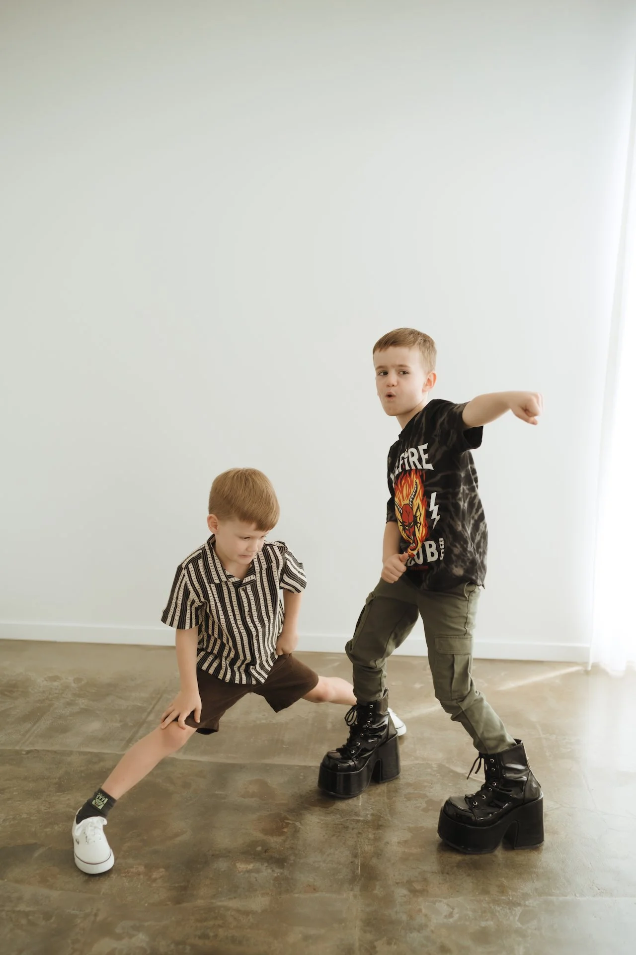 Two young boys playing indoors, one wearing large platform boots, with a plain wall in the background.