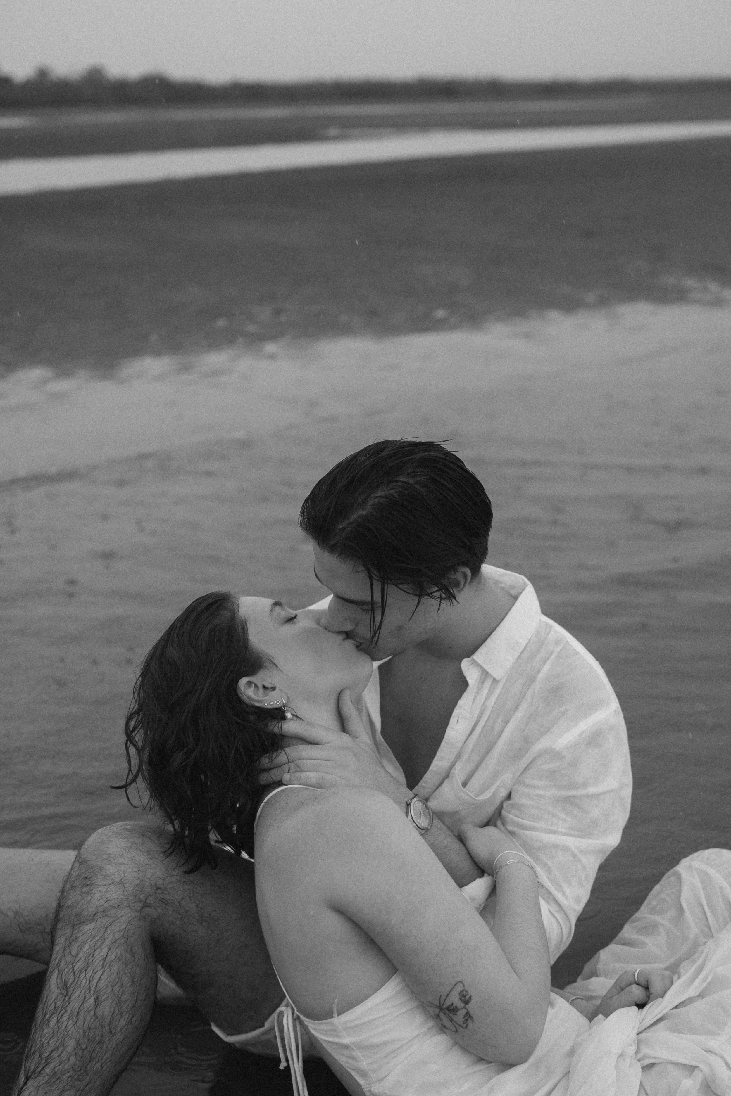 A black and white photo of a couple kissing by the beach, with the ocean in the background.