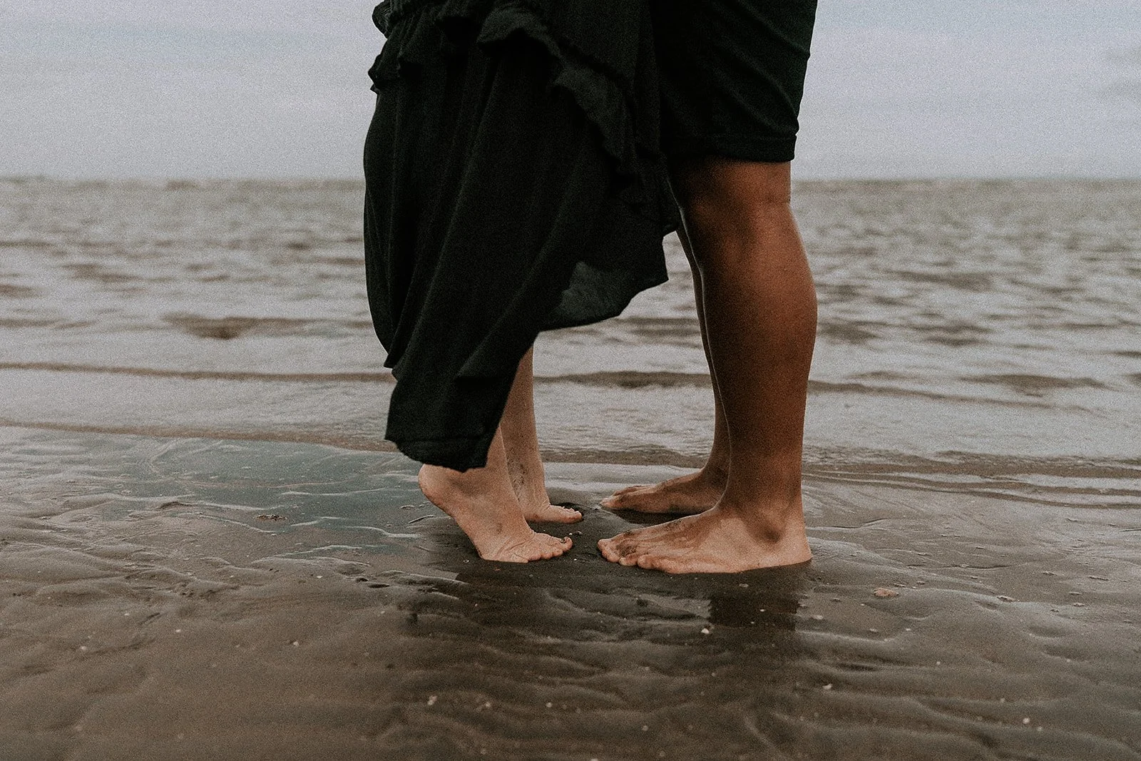 Two people, likely a child and an adult, standing barefoot on wet sand at the beach, with their feet close together and the ocean in the background.