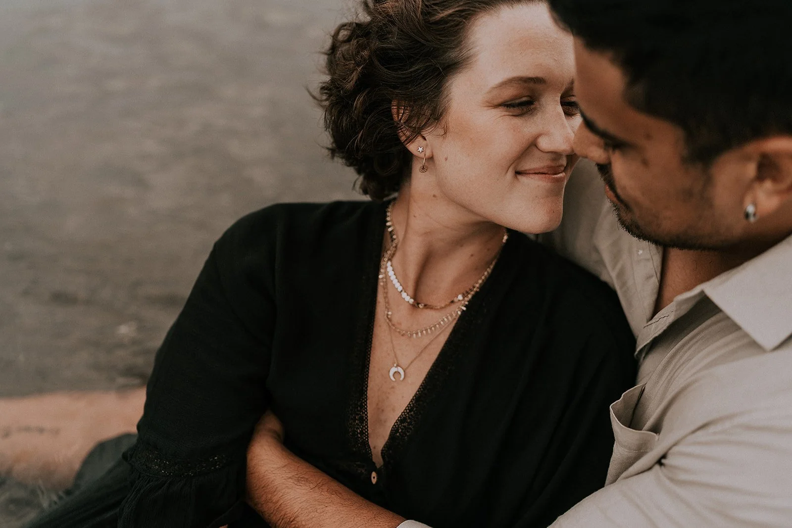 A woman and a man are close to each other, smiling with almost touching faces. The woman has short, curly hair and is wearing layered necklaces and earrings. The man has dark curly hair, a beard, and is wearing earrings and a light-colored shirt. The