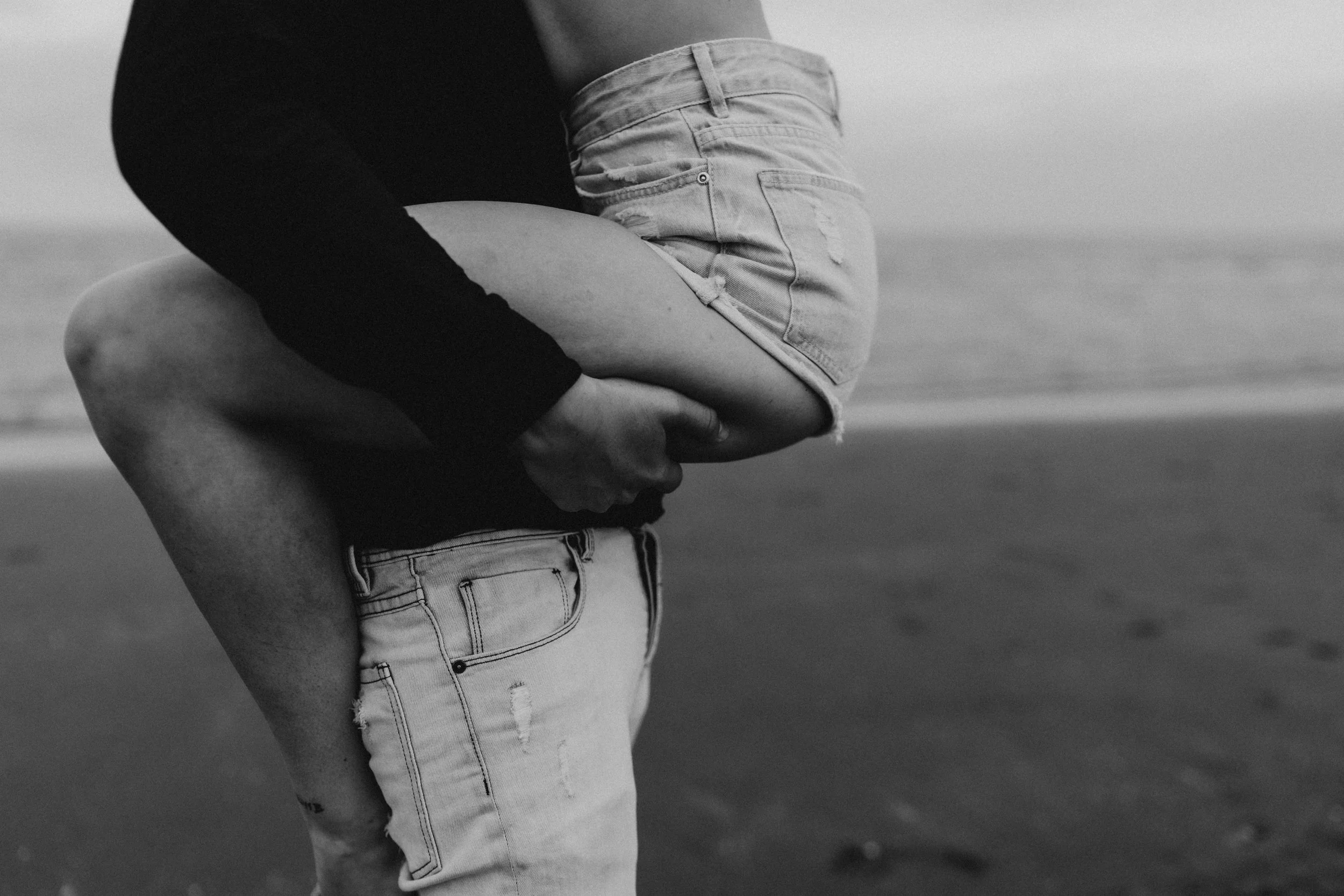 A couple on a beach, with the man holding the woman as she wraps her arms around his neck, in black and white.