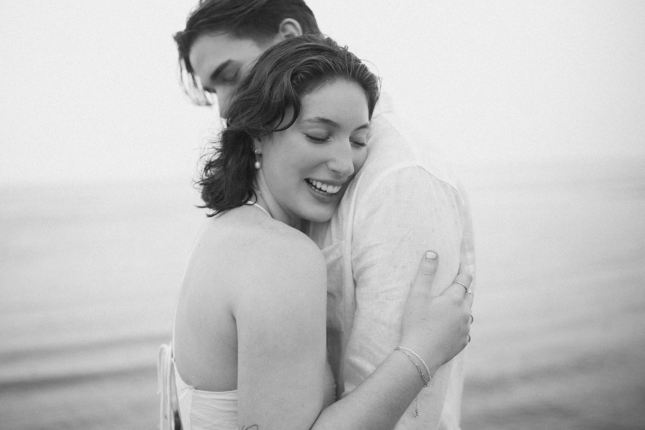 A black-and-white photo of a woman and man hugging each other at the beach, with the woman's eyes closed and smiling.