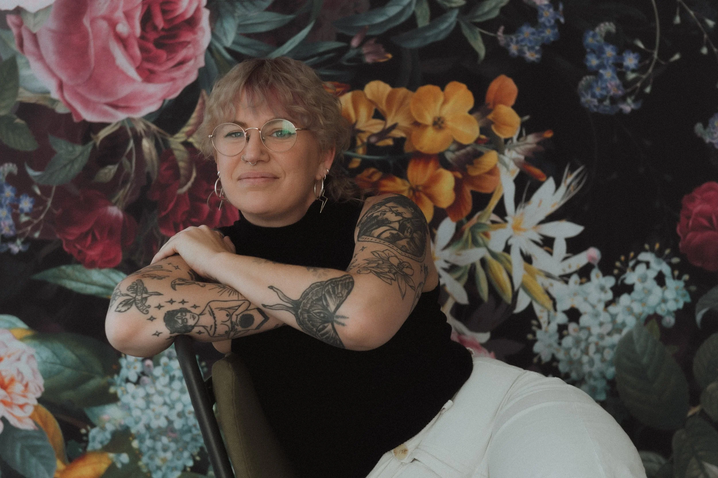 Person with short hair and glasses, wearing a black top and white pants, poses with tattooed arms against a vibrant floral background.