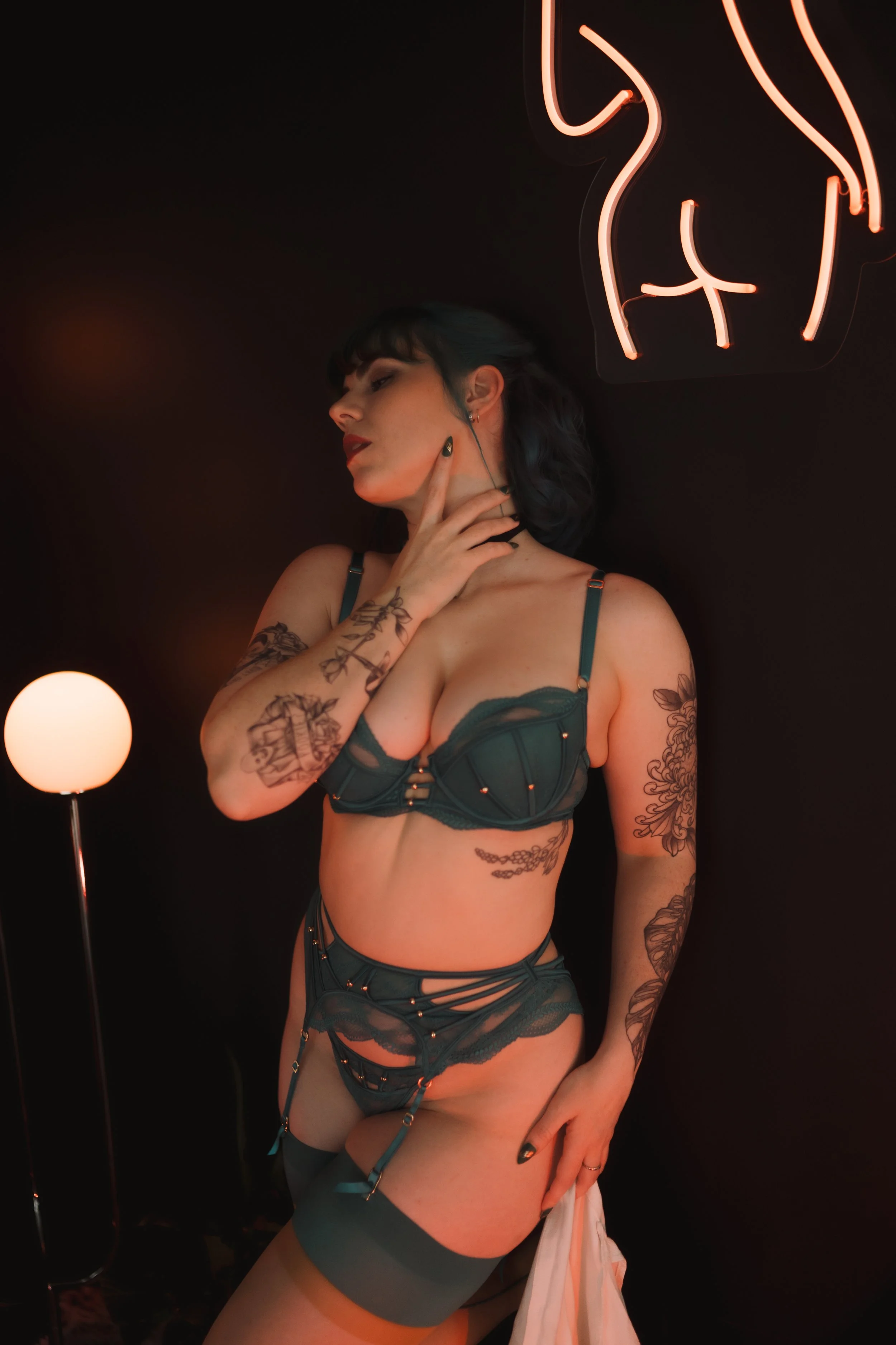 Person wearing green lingerie with tattoos, standing near a glowing neon sign and round lamp against a black background.