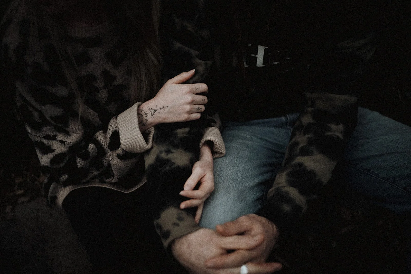 Two people sitting closely, one wearing a patterned jacket with tattoos on their wrist and hand, and the other in a dark patterned jacket with blue jeans, holding hands.