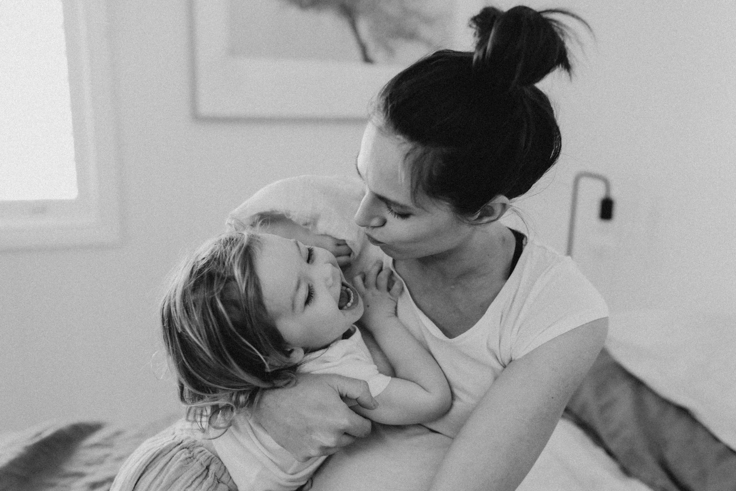 Black and white photo of a woman holding and kissing a smiling child in a cozy setting.