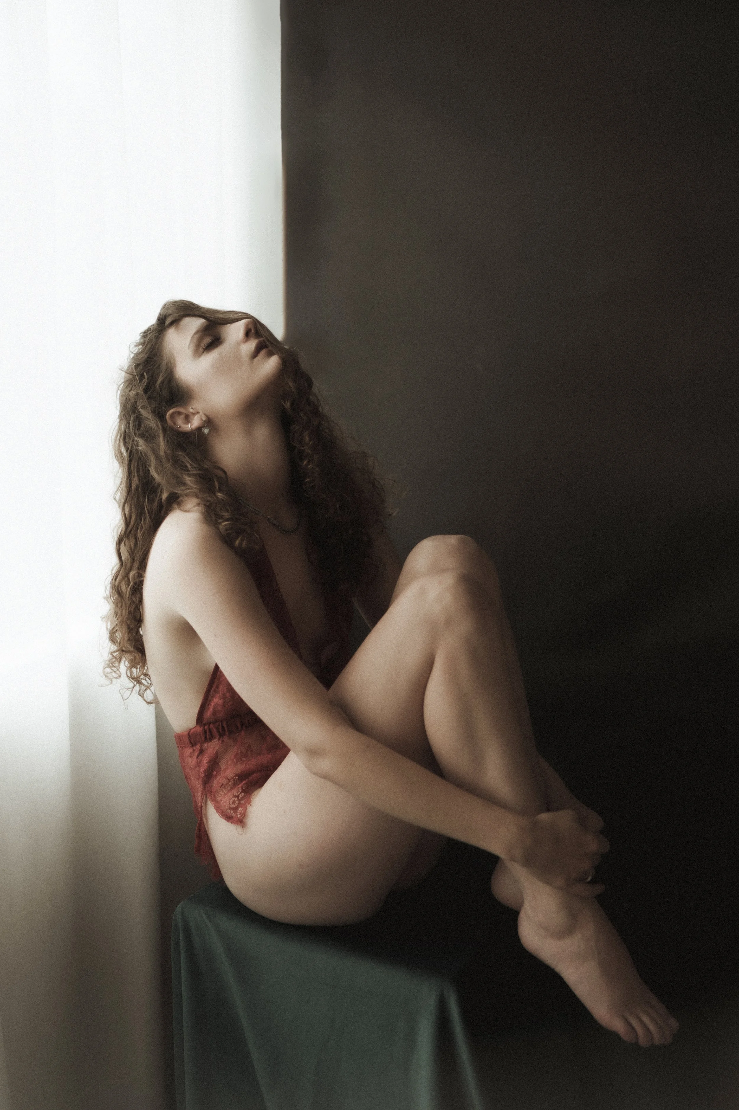 A woman sitting on a green stool, wearing a red lace bodysuit with her knees drawn to her chest. She has curly hair and is leaning back with her eyes closed, next to a bright window.