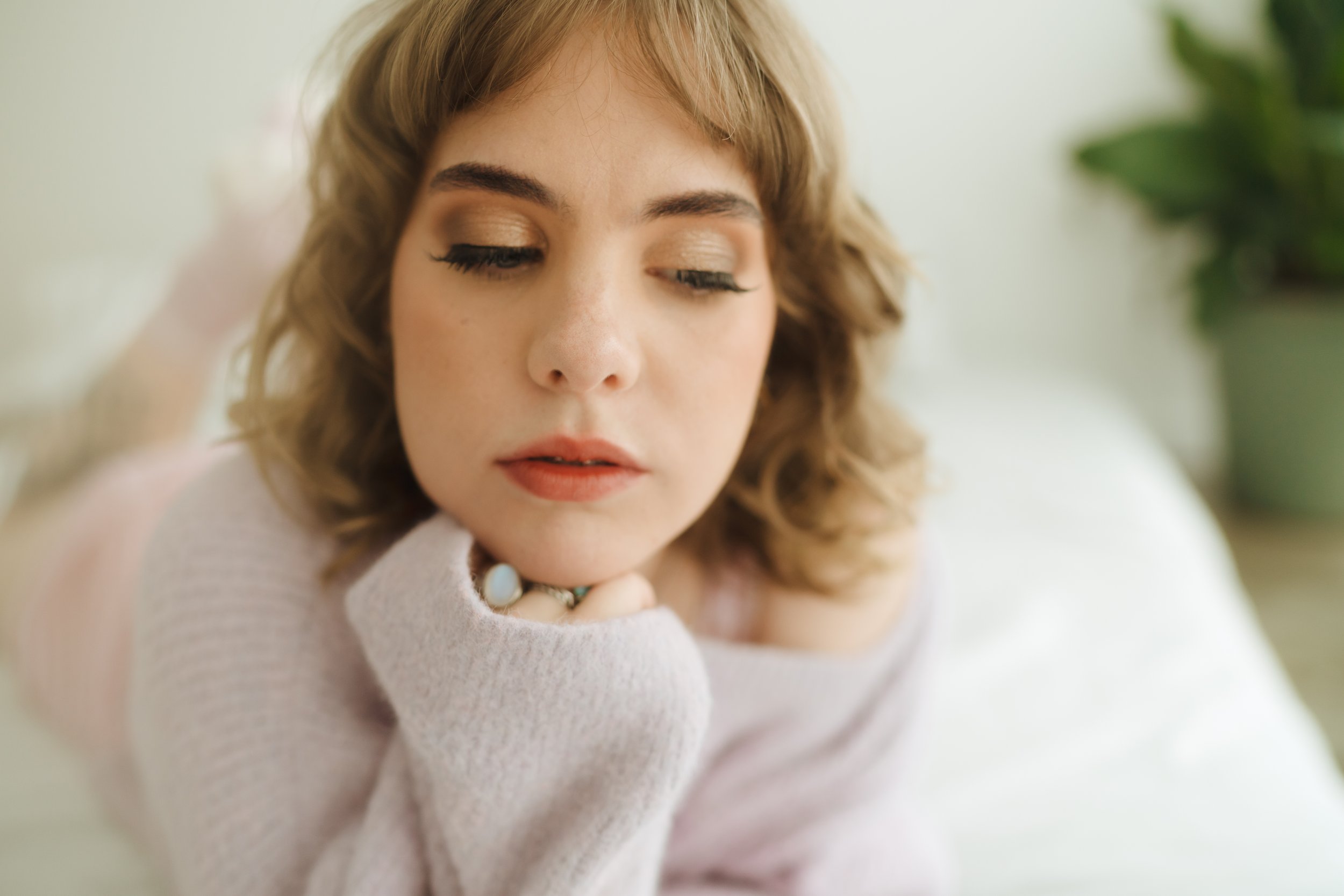 Woman with curly hair resting her chin on her hand, wearing a lavender sweater, eyes closed, indoor setting, plant in background.