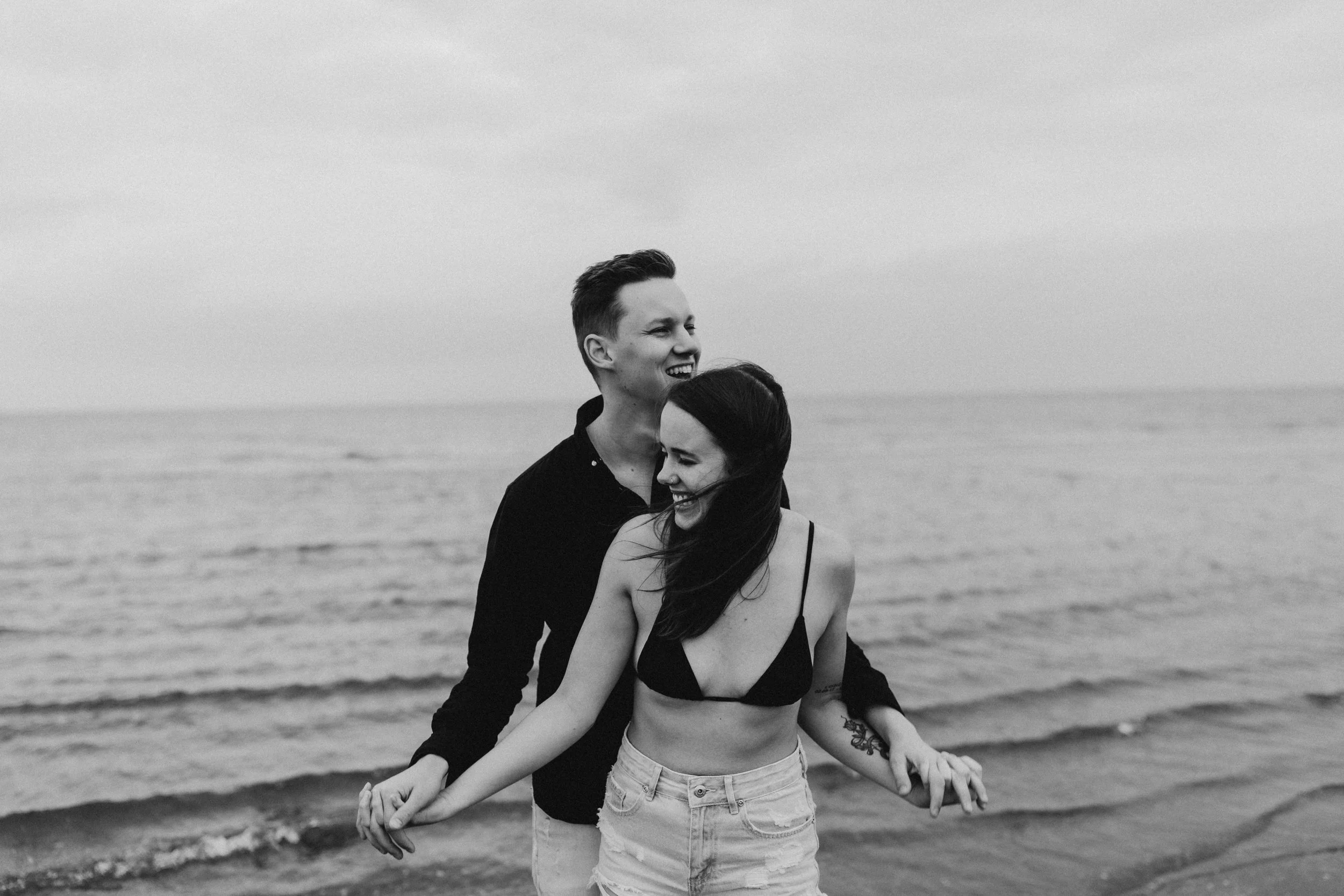 A young couple smiling and holding hands on the beach with the ocean in the background.