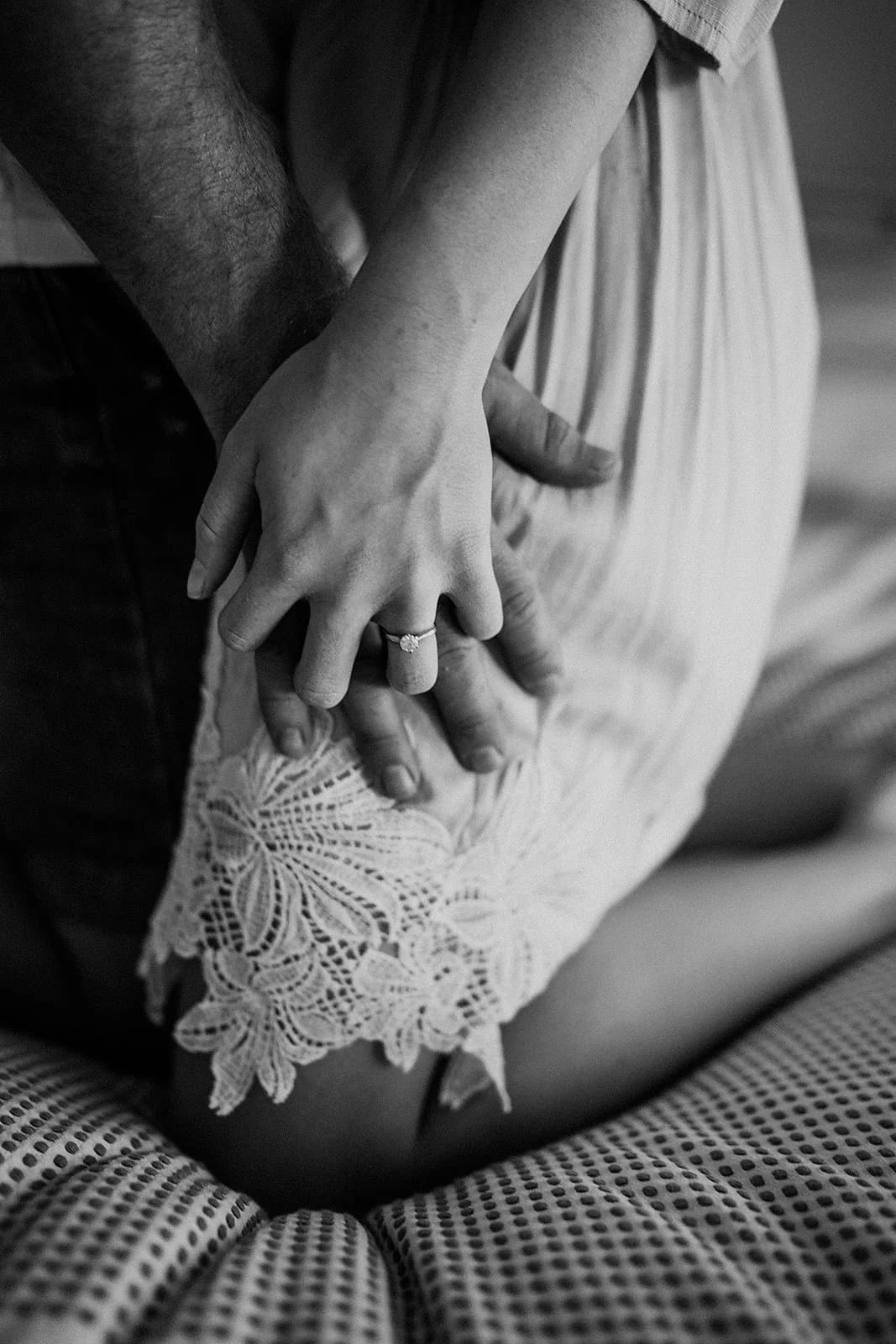 A couple showing their hands with a wedding ring, sitting close together on a textured surface. The woman's hand rests on the man's hand, with a floral lace dress visible.