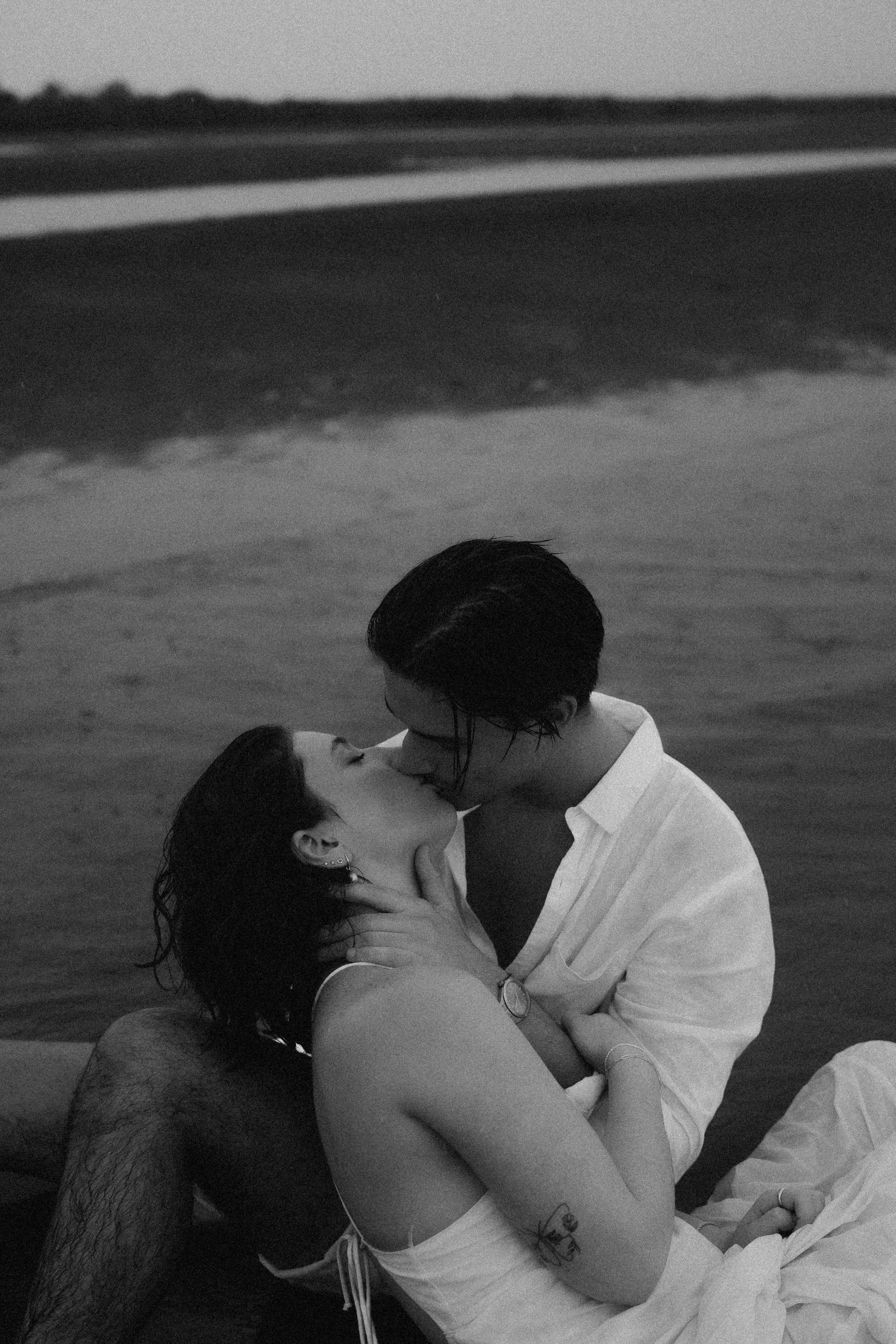 Black and white photo of a couple kissing by the water.
