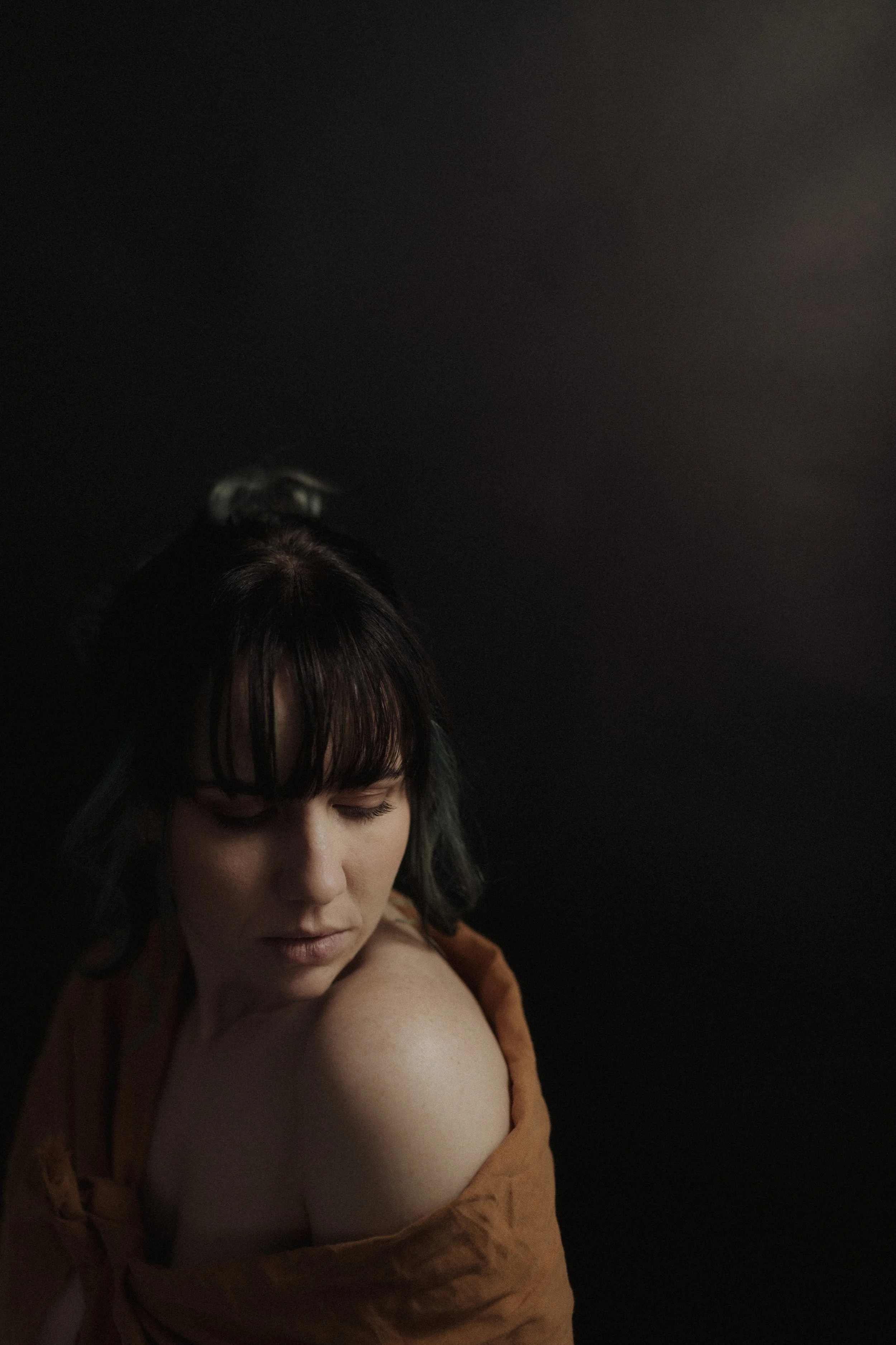 Portrait of a woman with dark hair and bangs, wrapped in a brown fabric, looking down with a dark background.