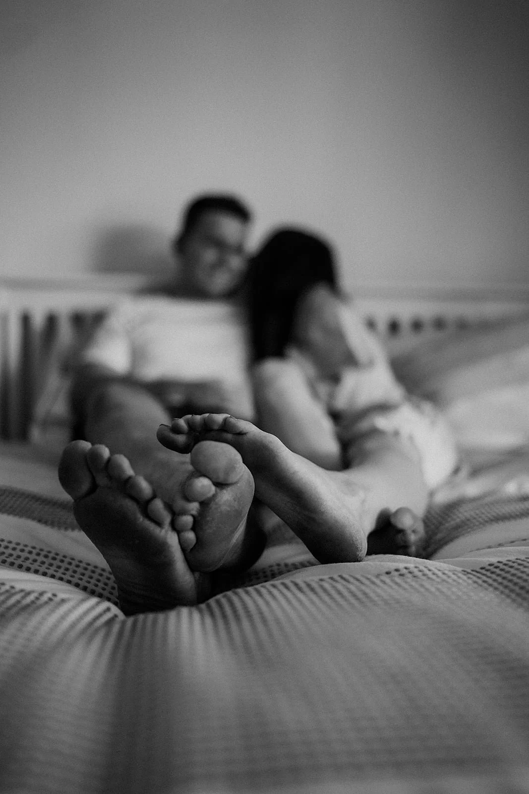 A black and white photo of a couple sitting on a bed, focusing on their feet in the foreground, with the couple blurred in the background.