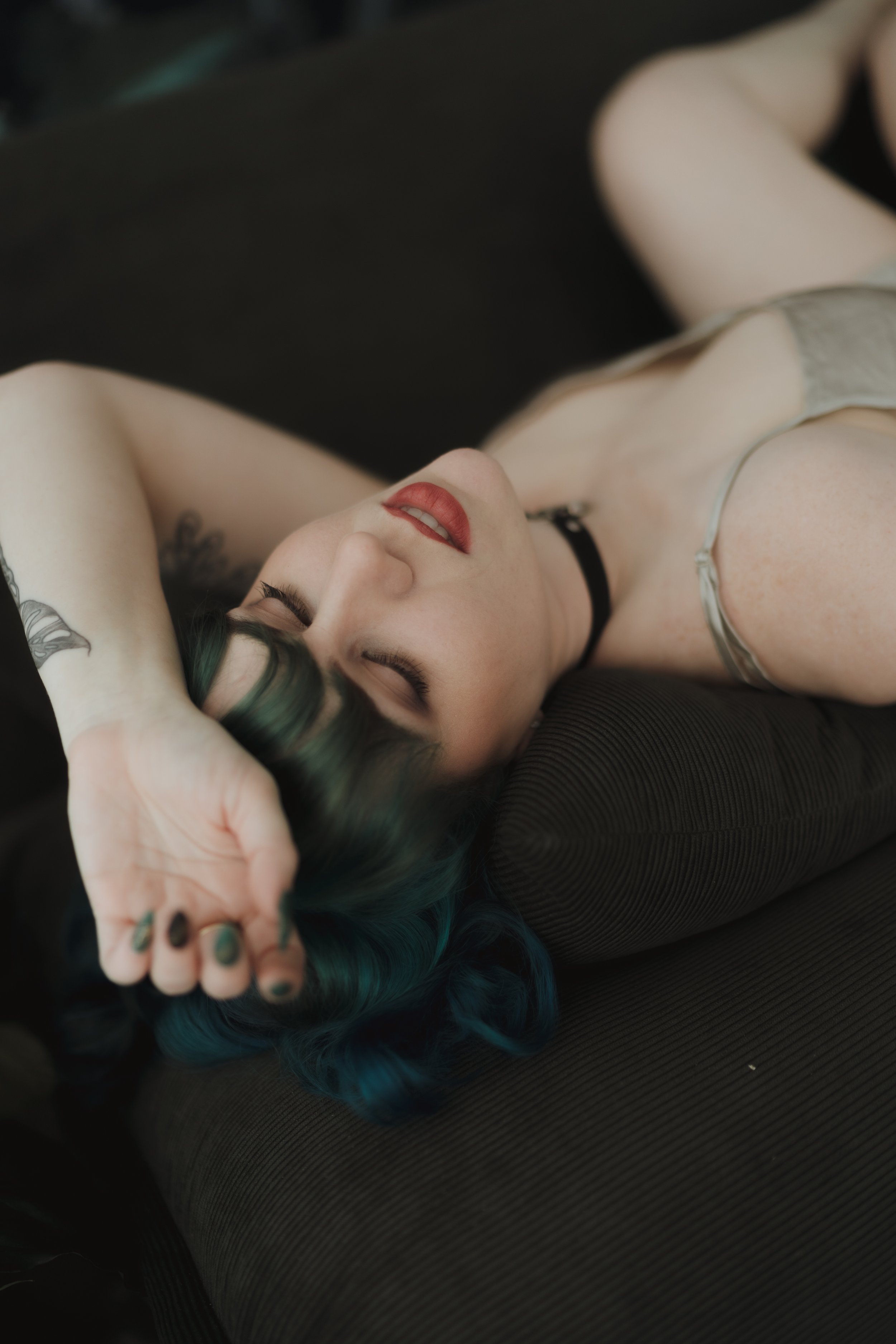 Person with blue hair and red lipstick lying on dark cushions, hand on forehead, wearing a black choker.