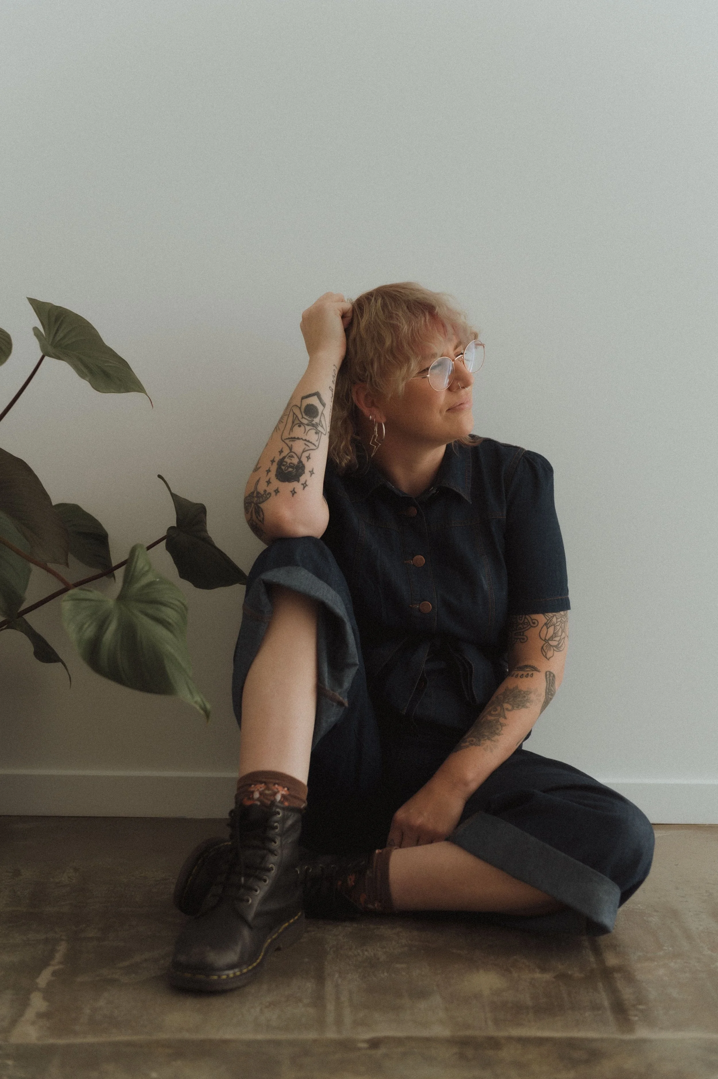 Person with tattoos sitting on the floor next to a potted plant, wearing glasses, a dark jumpsuit, and boots.