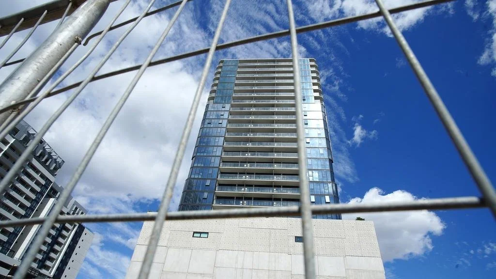 High-rise building view through metal fence, with bright blue sky and white clouds behind.