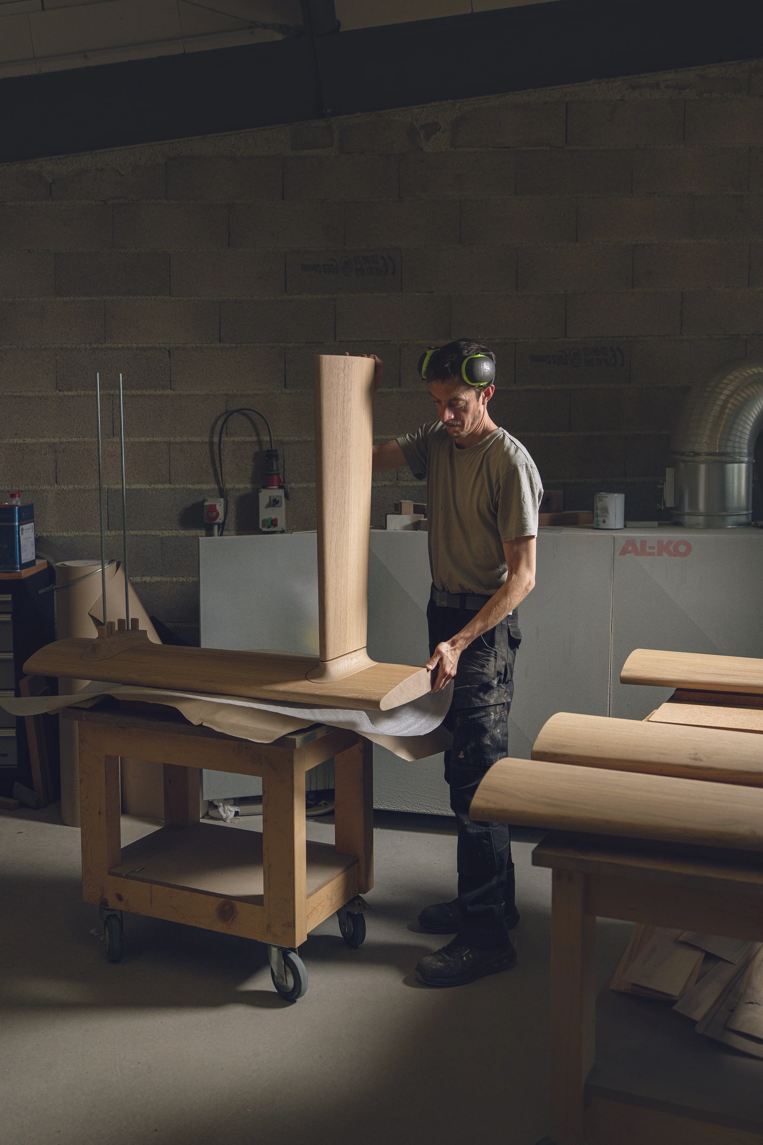 A man wearing safety glasses and ear protection works on a piece of wood in a woodworking shop with various tools and furniture in the background.