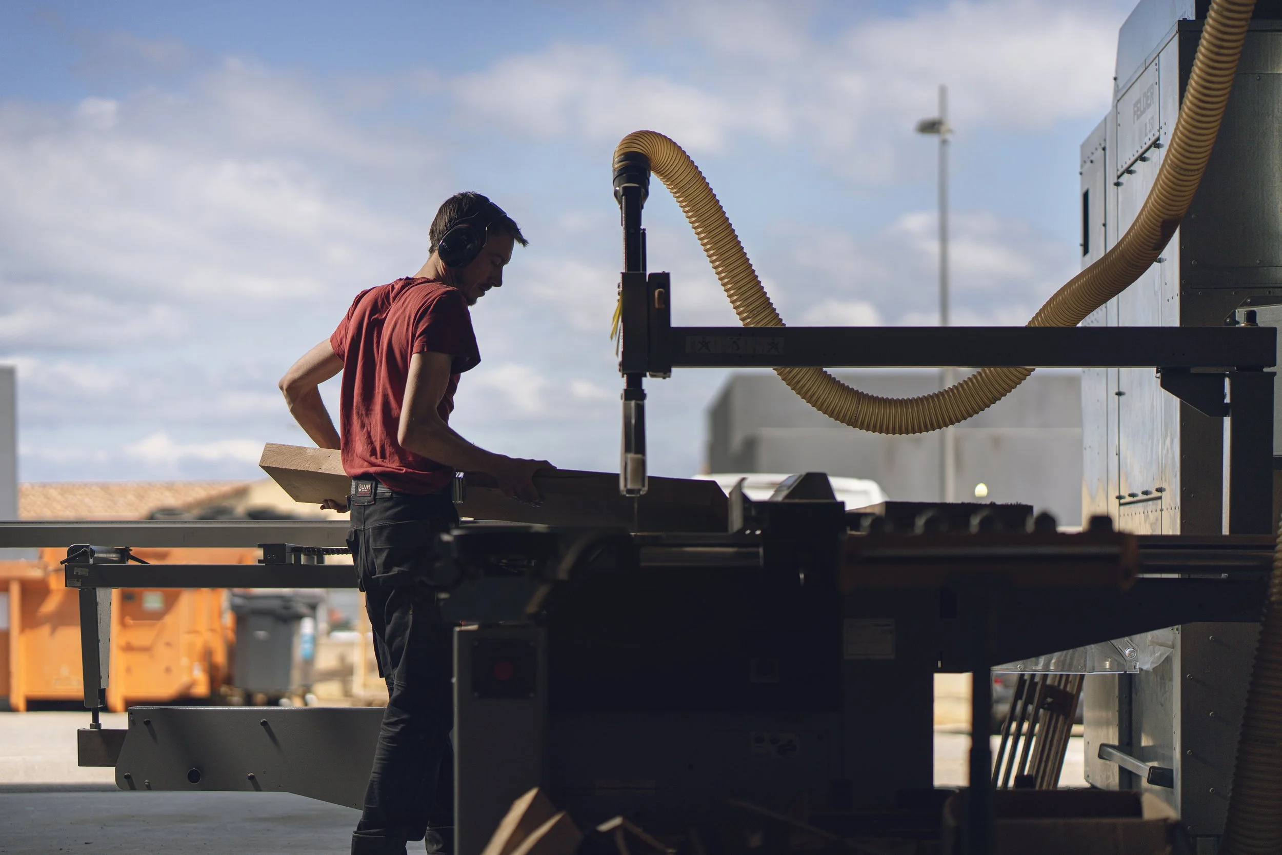 A man wearing a red shirt and headphones is working with woodworking equipment outdoors under a blue sky with clouds.