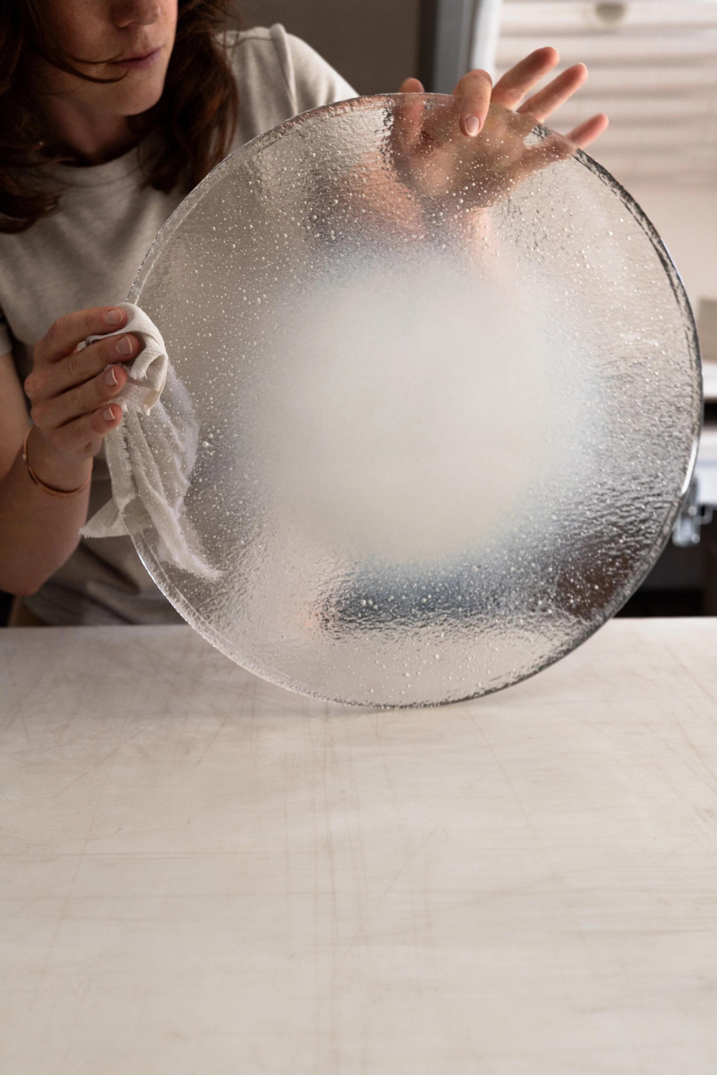 A person cleaning a round glass plate with a white cloth, with water droplets and fog on the glass.