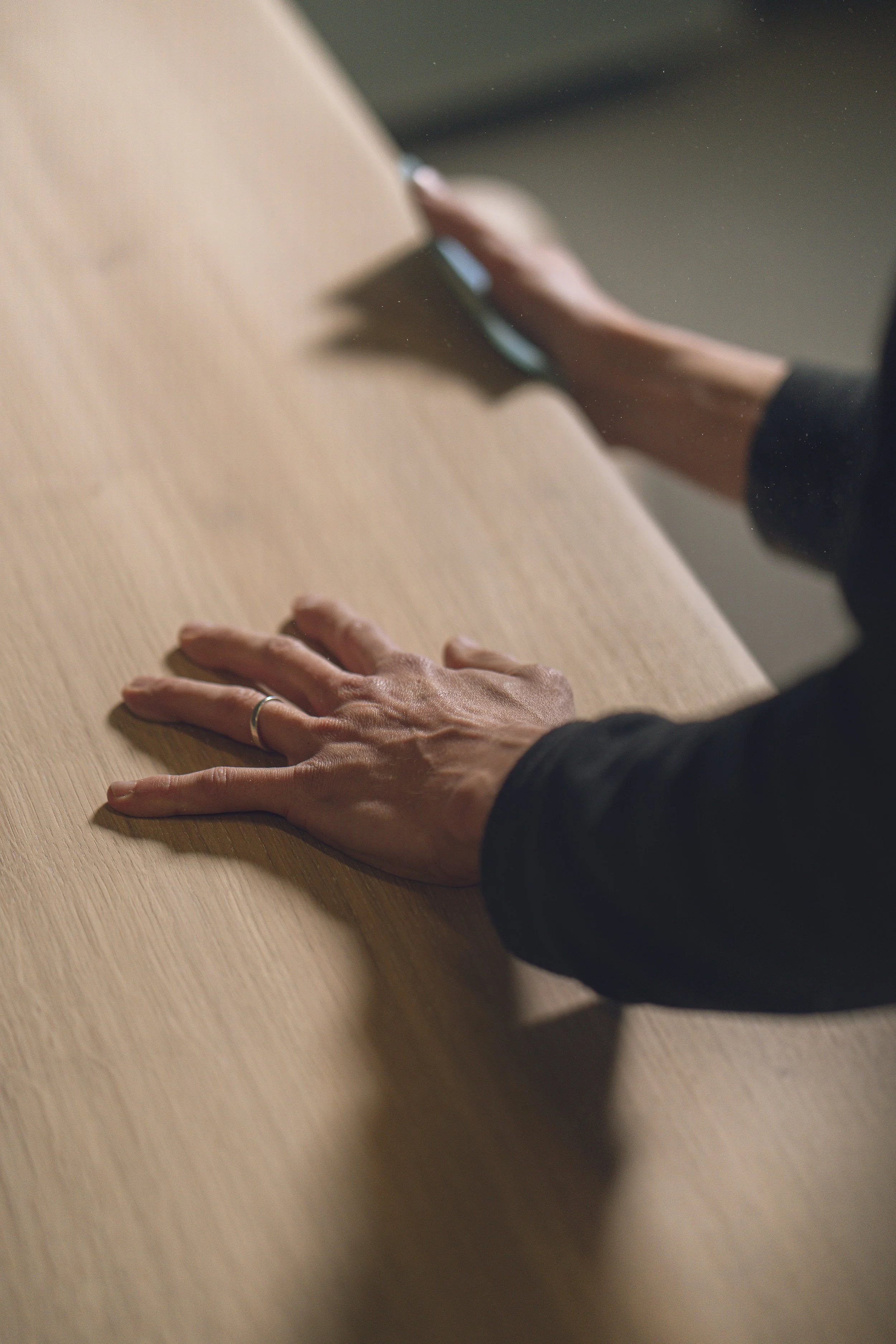 A person's hands resting on a wooden table, with one hand having a ring on the finger and the other hand reaching towards the table, with a slight shadow cast on the surface.
