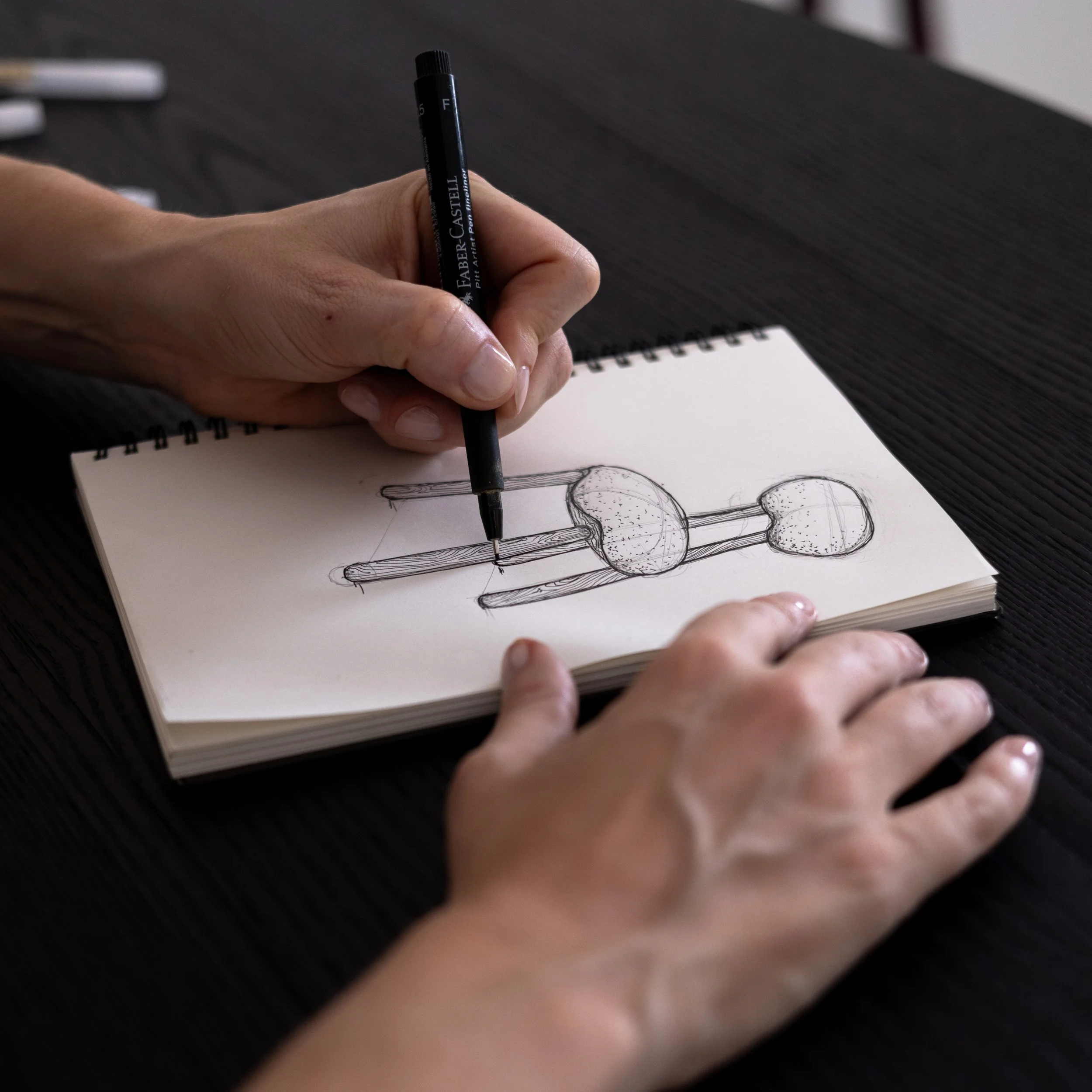 A person holding a black pen with the label 'Faber-Castell' drawing in a spiral sketchbook on a dark surface.