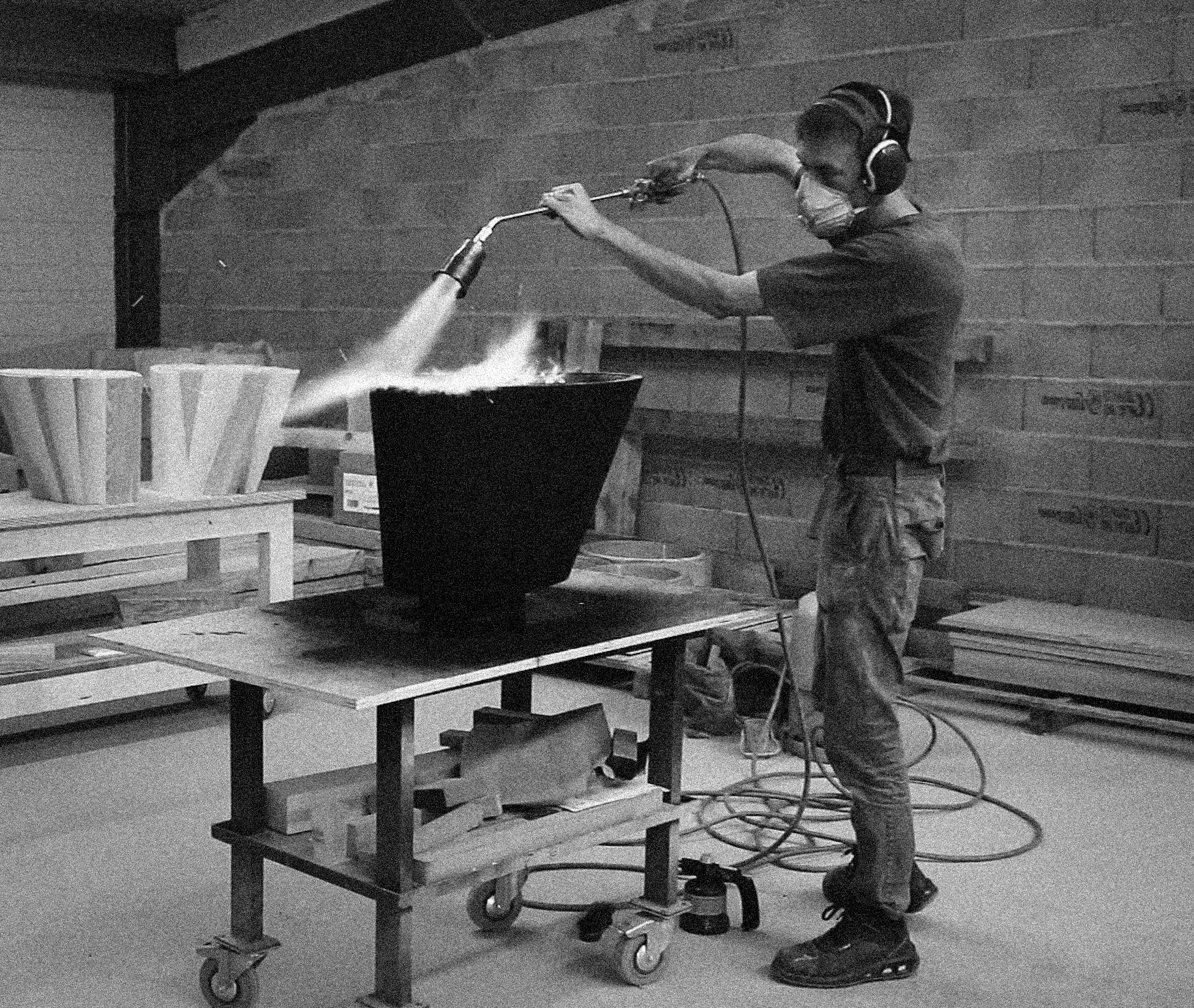 A person wearing a mask, headphones, and safety shoes operating a spray machine to paint a large metal container in an industrial workshop with wooden pallets.