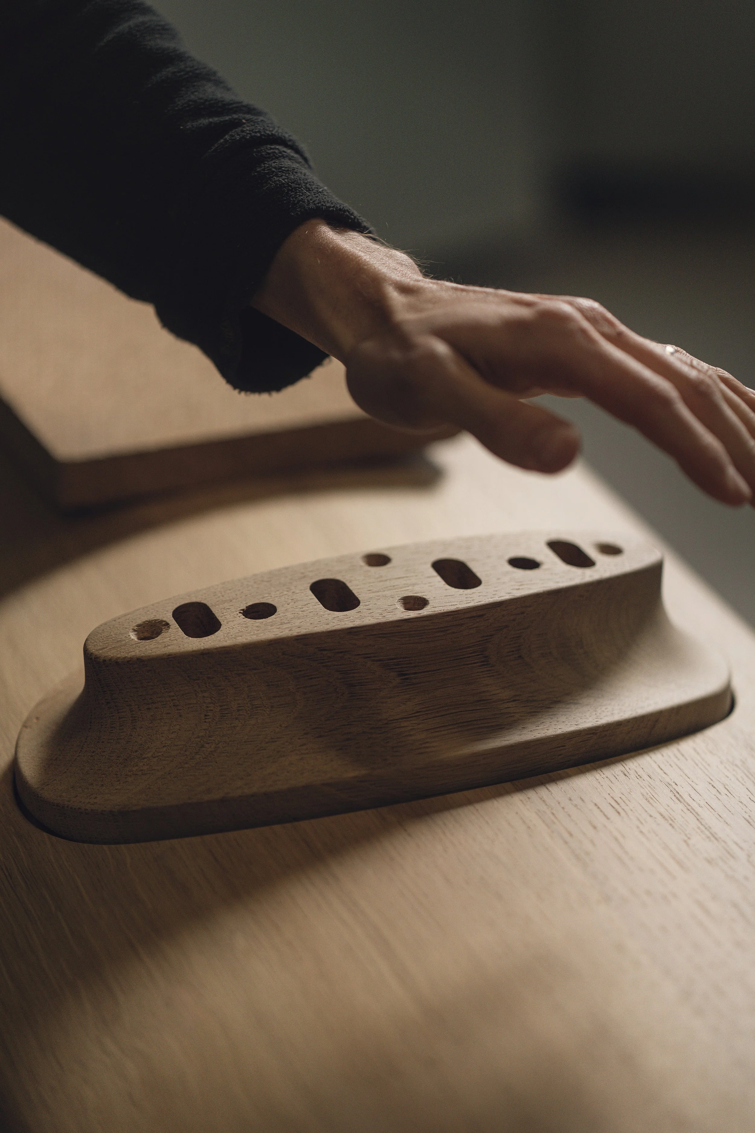 Close-up of a person's hand reaching out towards a wooden object with holes, on a wooden surface, with a cardboard box nearby.